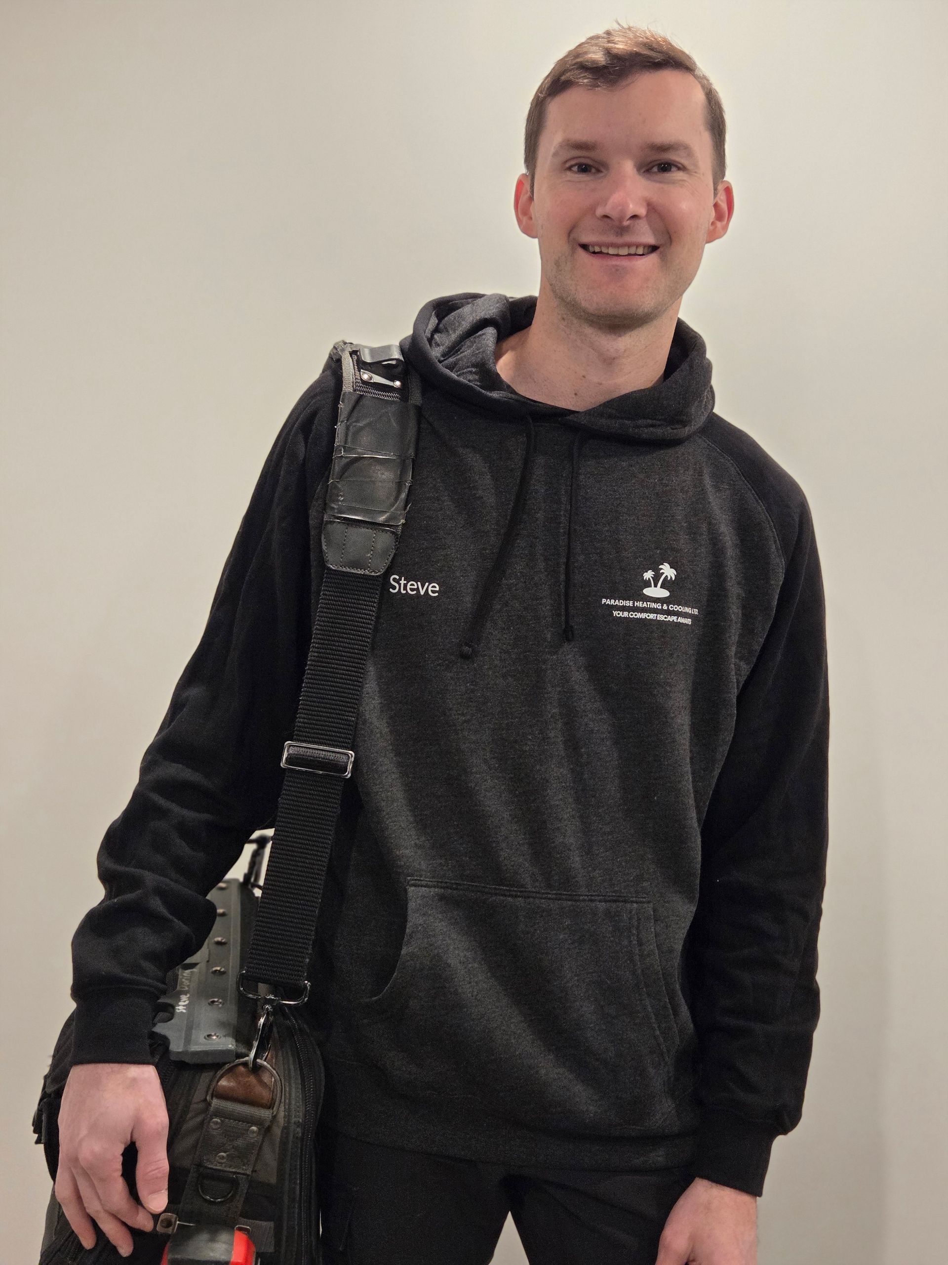 Man smiling, wearing a black hoodie and carrying a bag, standing against a light gray wall.