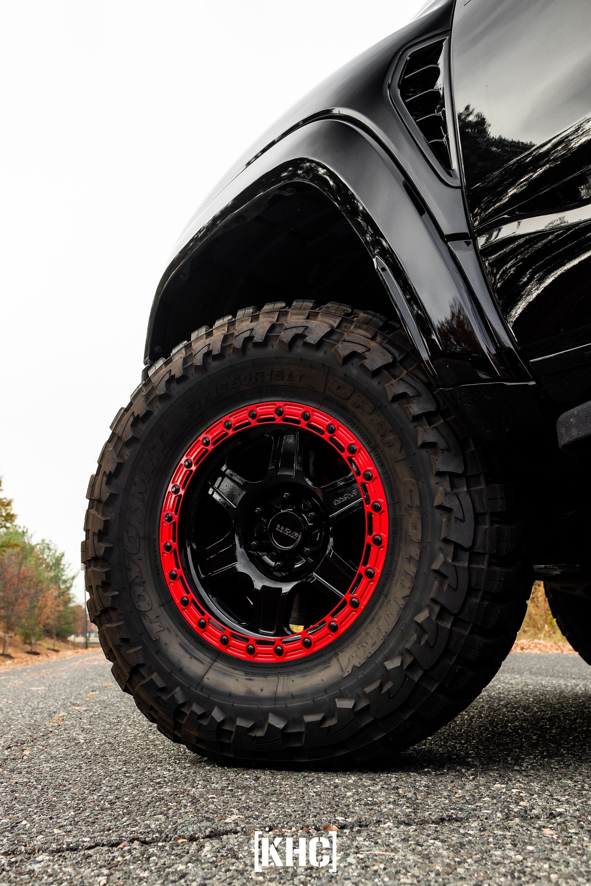Black off-road tire with red rim on asphalt road; vehicle partially visible against a cloudy sky.