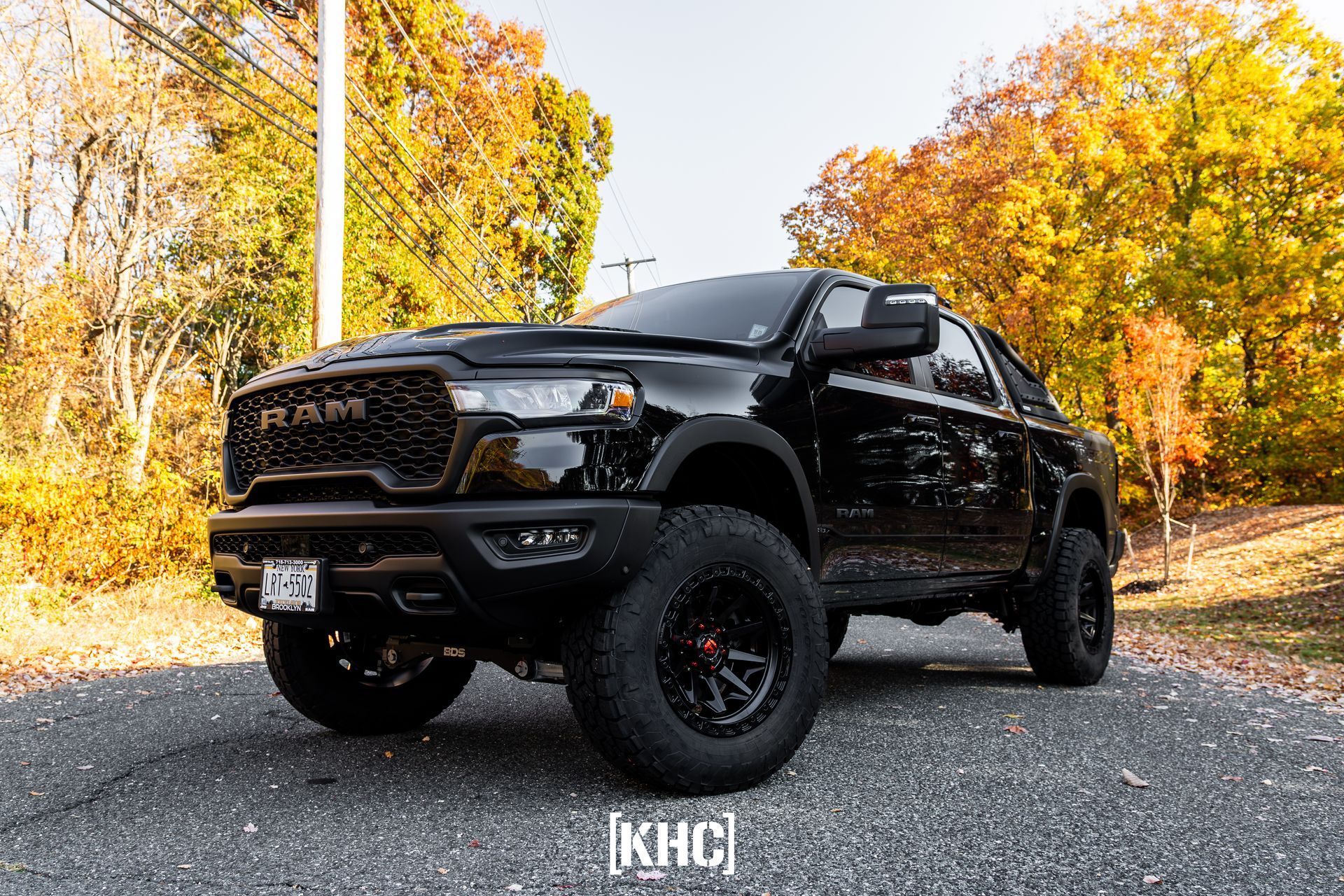 Black Ram TRX truck with off-road tires, parked on a road, fall foliage background.