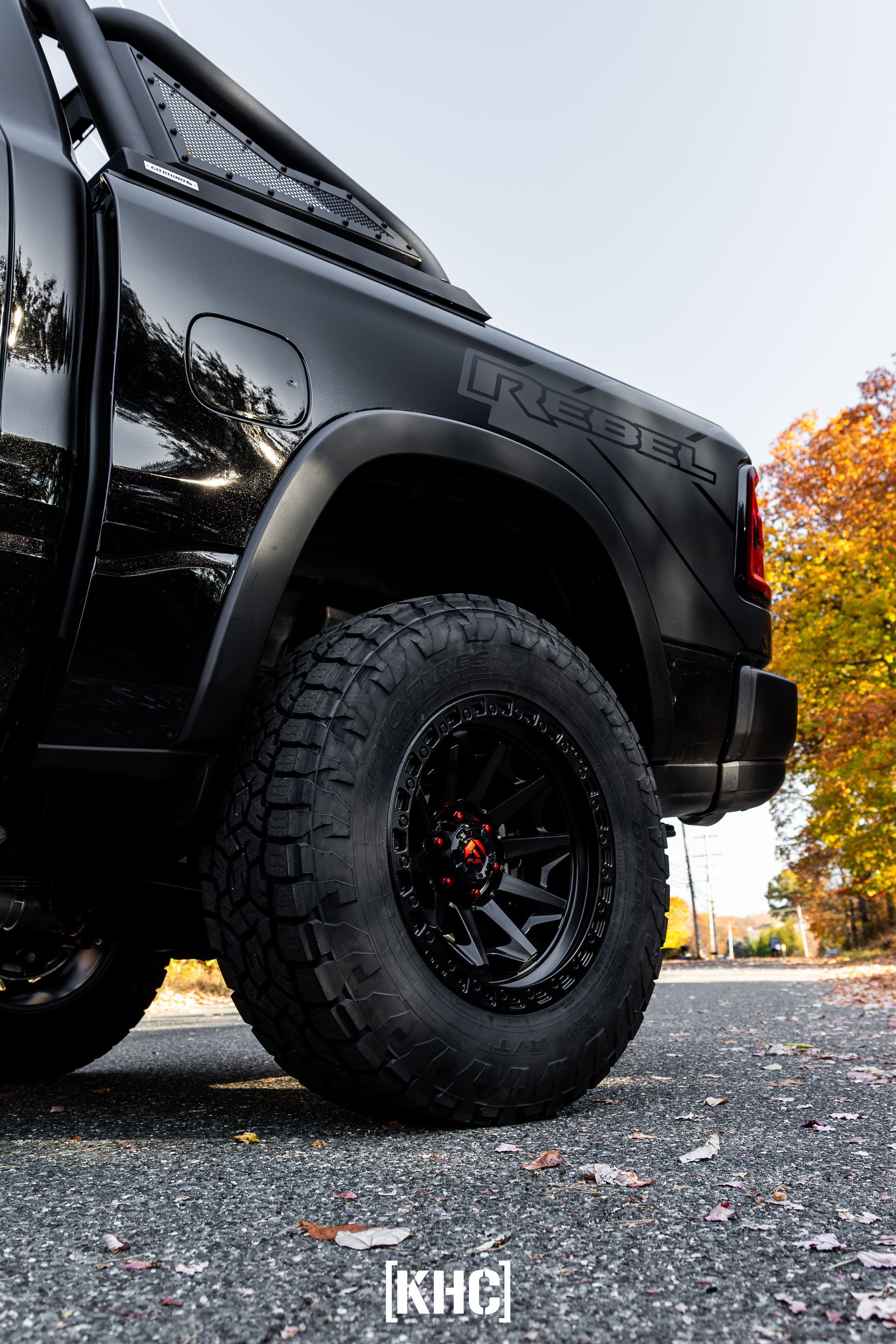 Black RAM TRX truck, rear wheel visible, parked on a road with fall foliage.