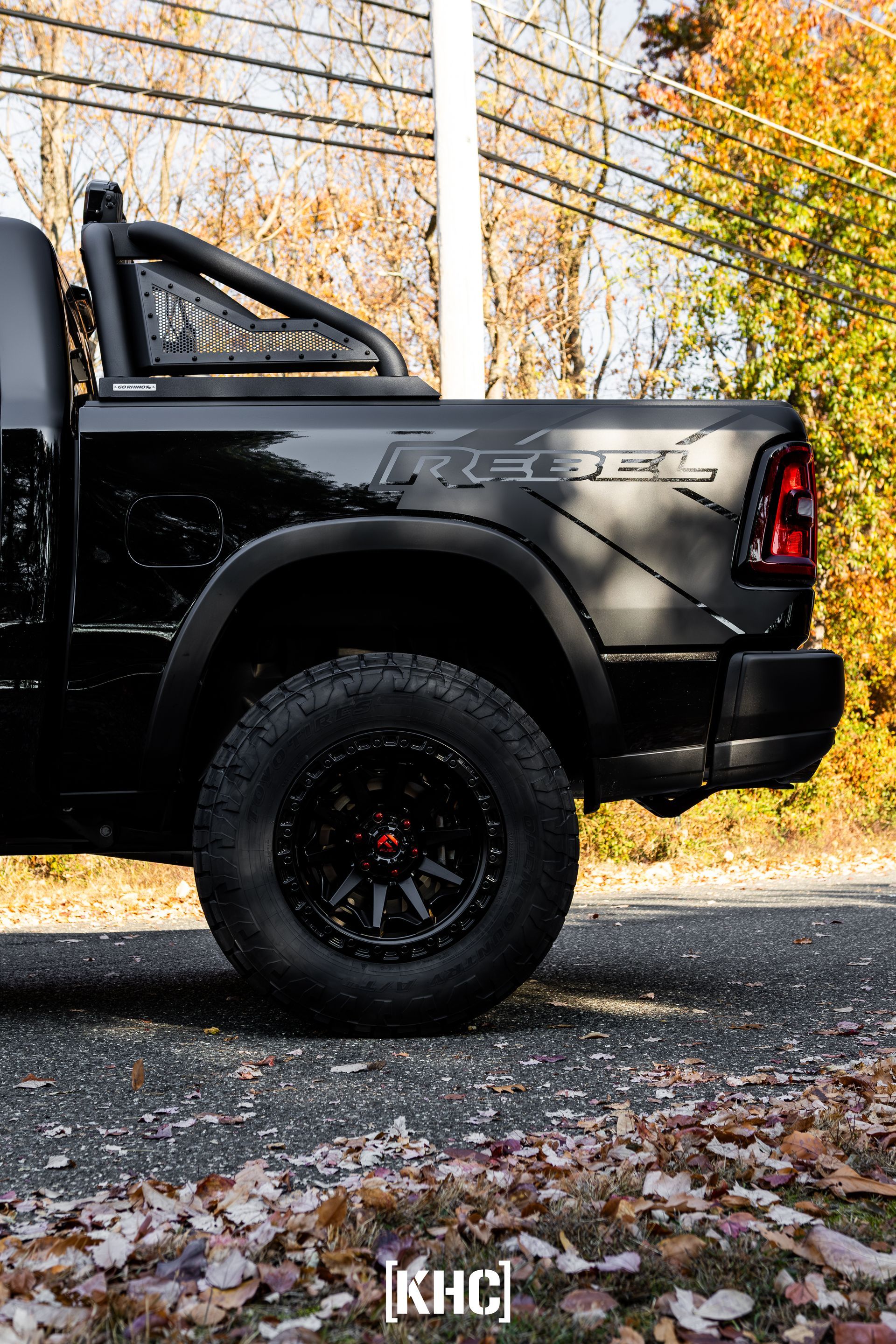 Black Ram Rebel truck with off-road wheels, roll bar, and Rebel decal, parked on a road with autumn leaves.