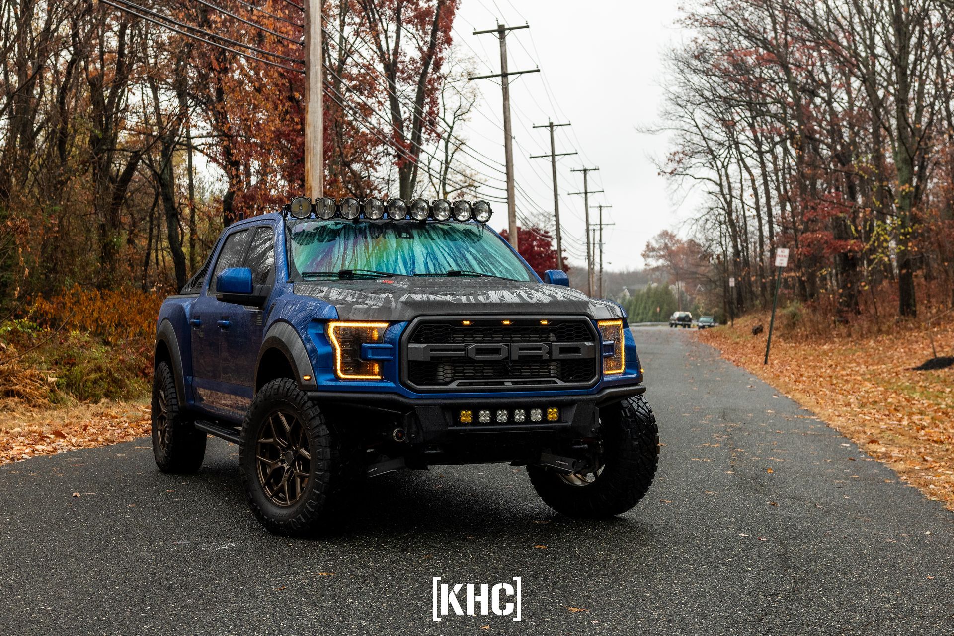 Blue Ford Raptor truck on a wet road, surrounded by autumn trees.  Lights mounted on the roof and front bumper.