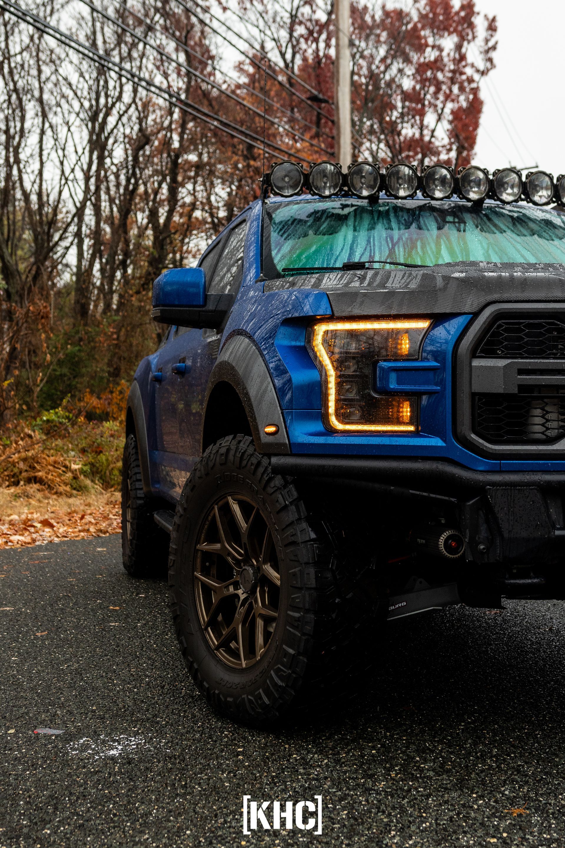 Blue Ford Raptor truck with off-road lights, parked on a wet road, in front of trees.