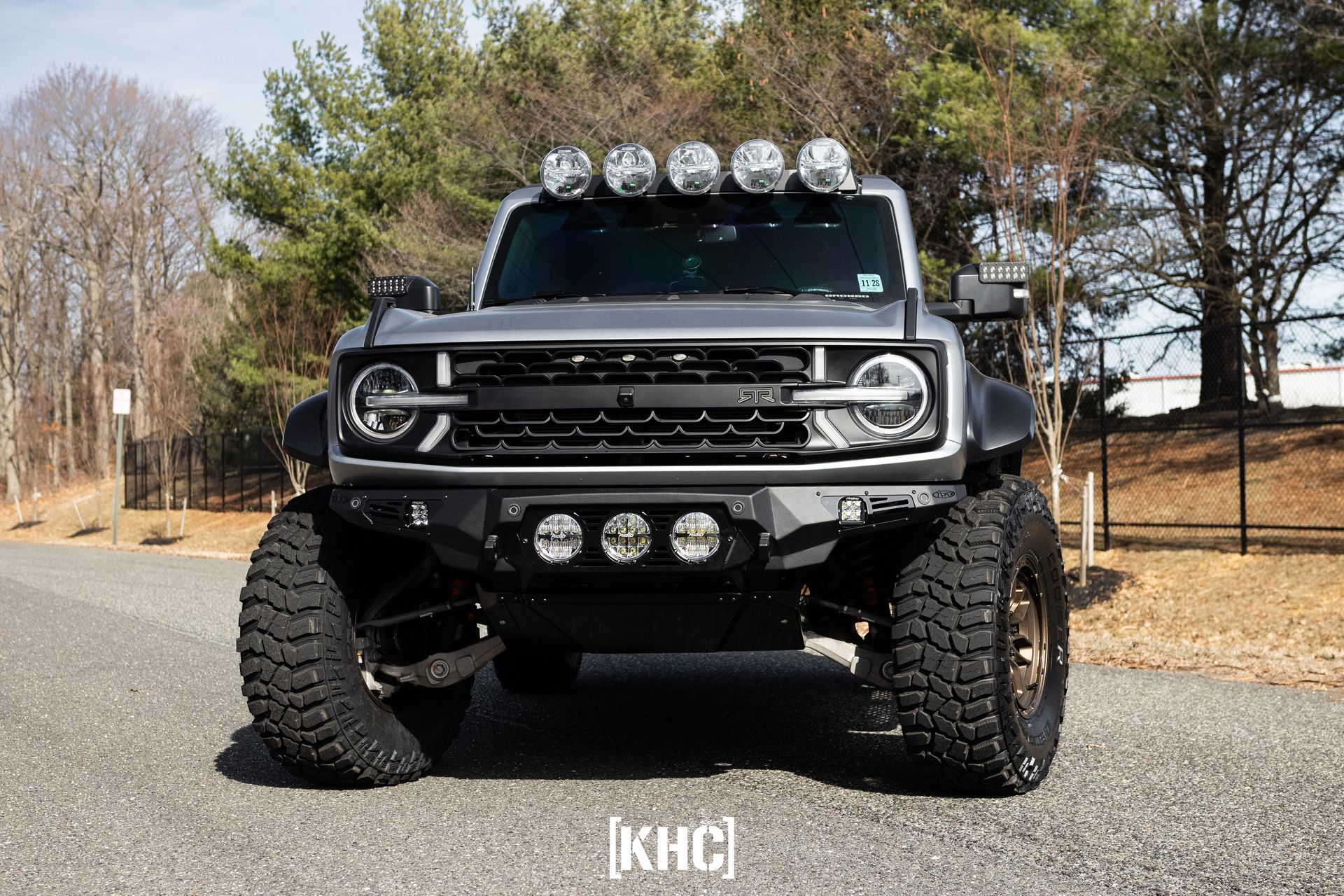 Gray, lifted Ford Bronco with off-road modifications, light bars, and large tires, parked on a road.