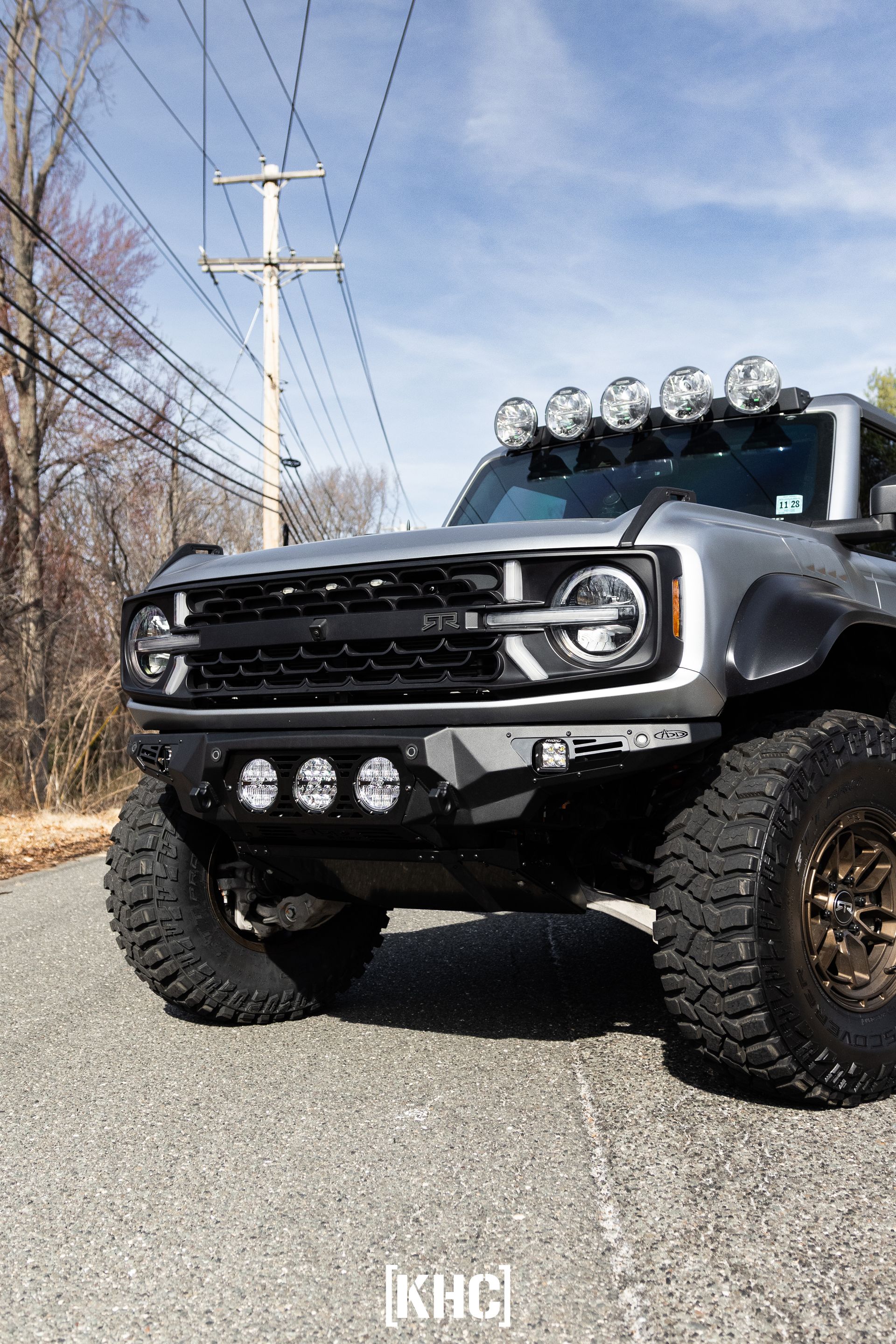 Silver Ford Bronco with off-road modifications, including a light bar, parked on a road.