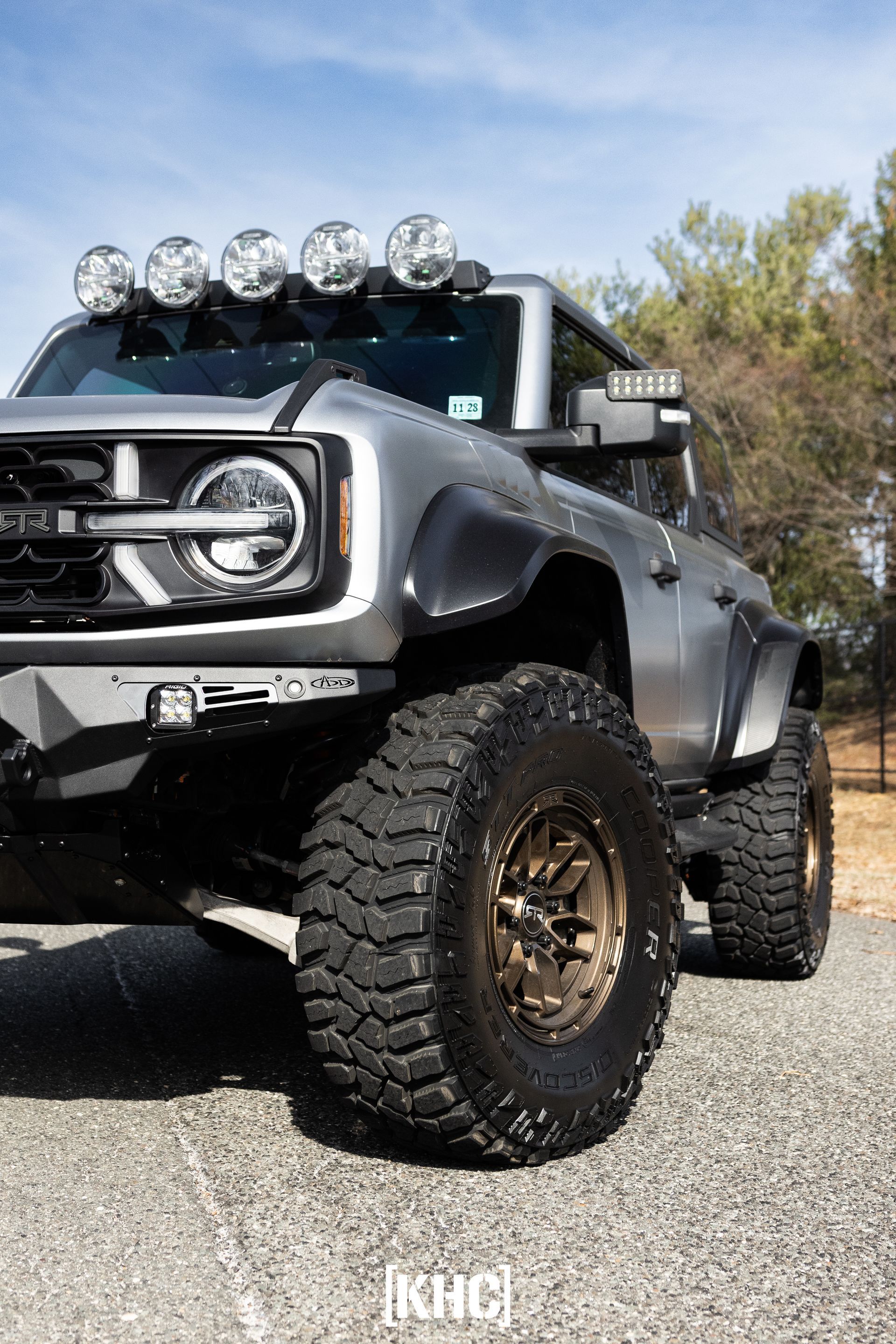 Gray Ford Bronco with oversized tires, roof lights, and custom bumper on asphalt.