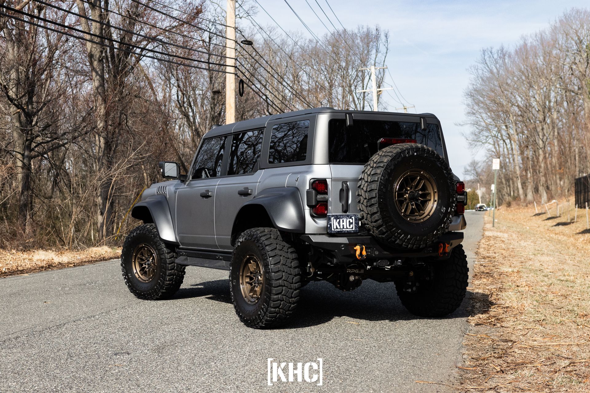 Gray Ford Bronco SUV on a gravel road, spare tire mounted, off-road wheels, surrounded by trees.