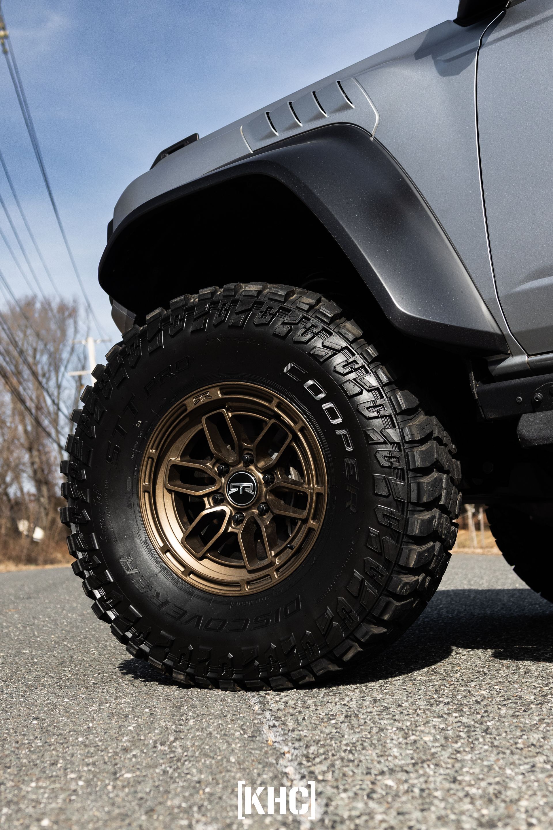 Close-up of a silver off-road vehicle's black tire and bronze rim on a paved road, under a blue sky.