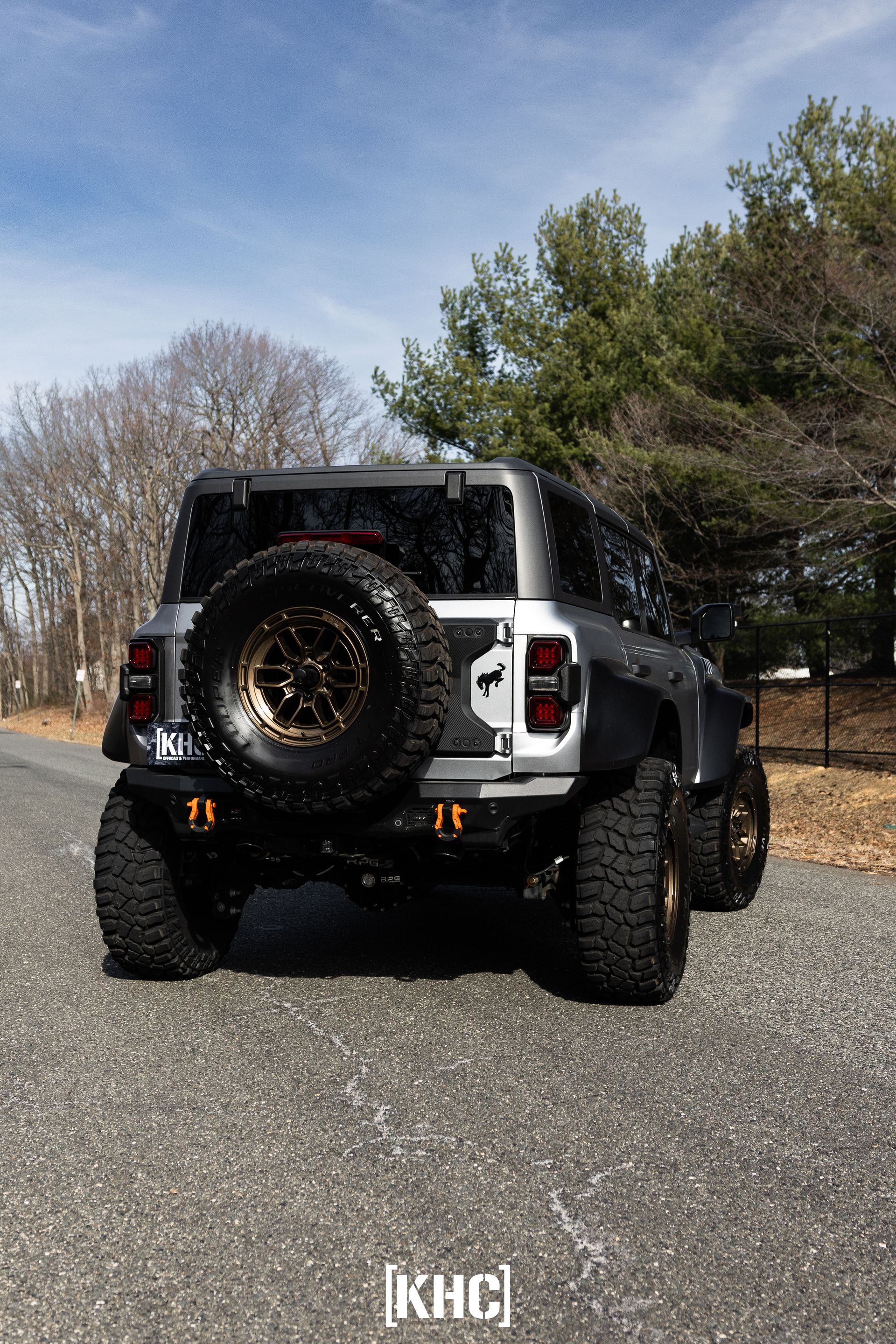 Gray modified Jeep with large tires on gravel road, sunny day.