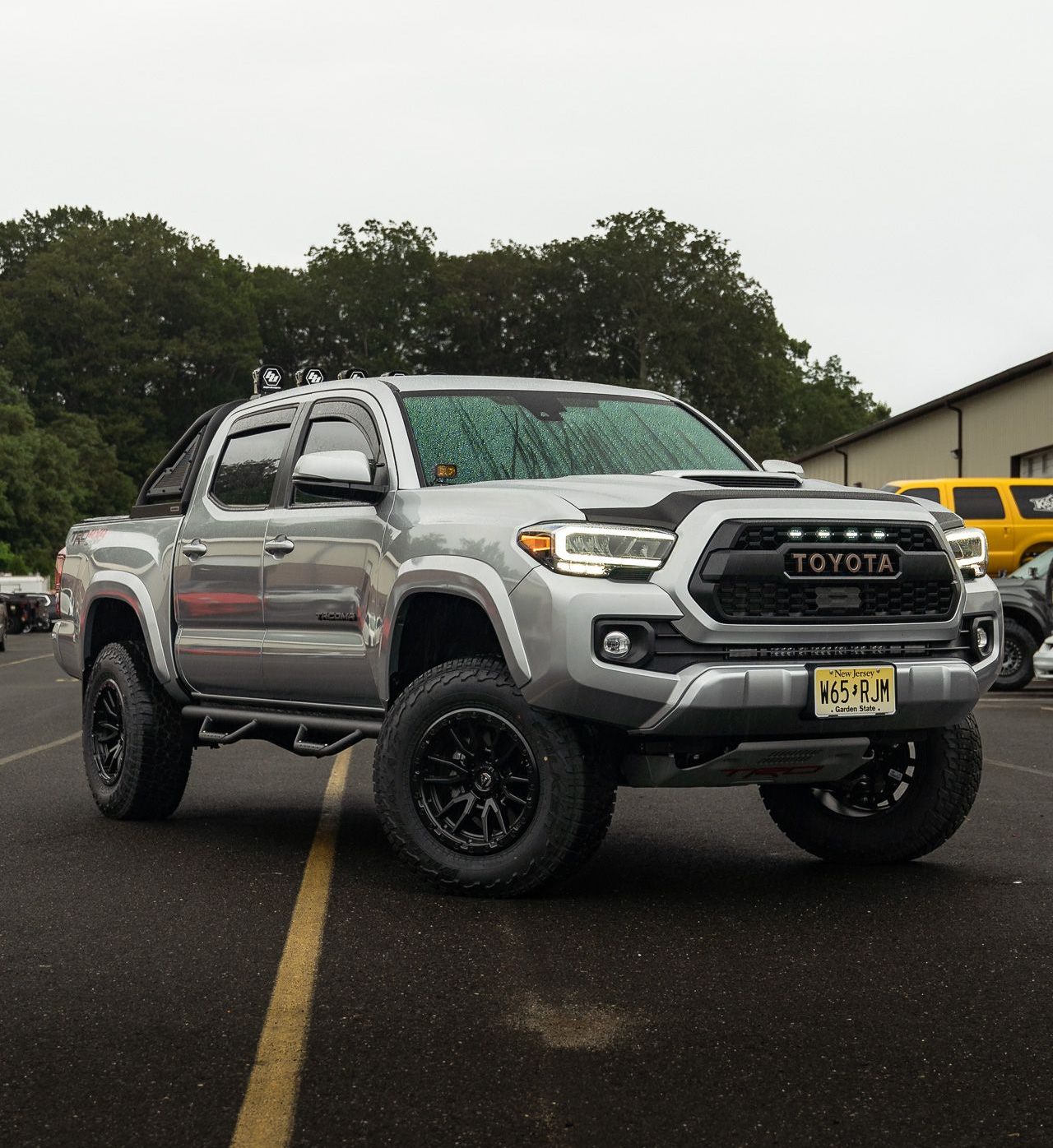 Silver Toyota Tacoma truck with black wheels parked on asphalt, cloudy day.