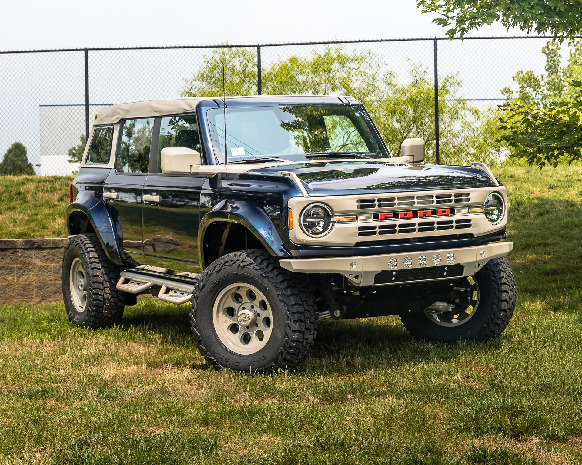 Blue and tan customized Ford Bronco parked on grass, next to a chain-link fence.