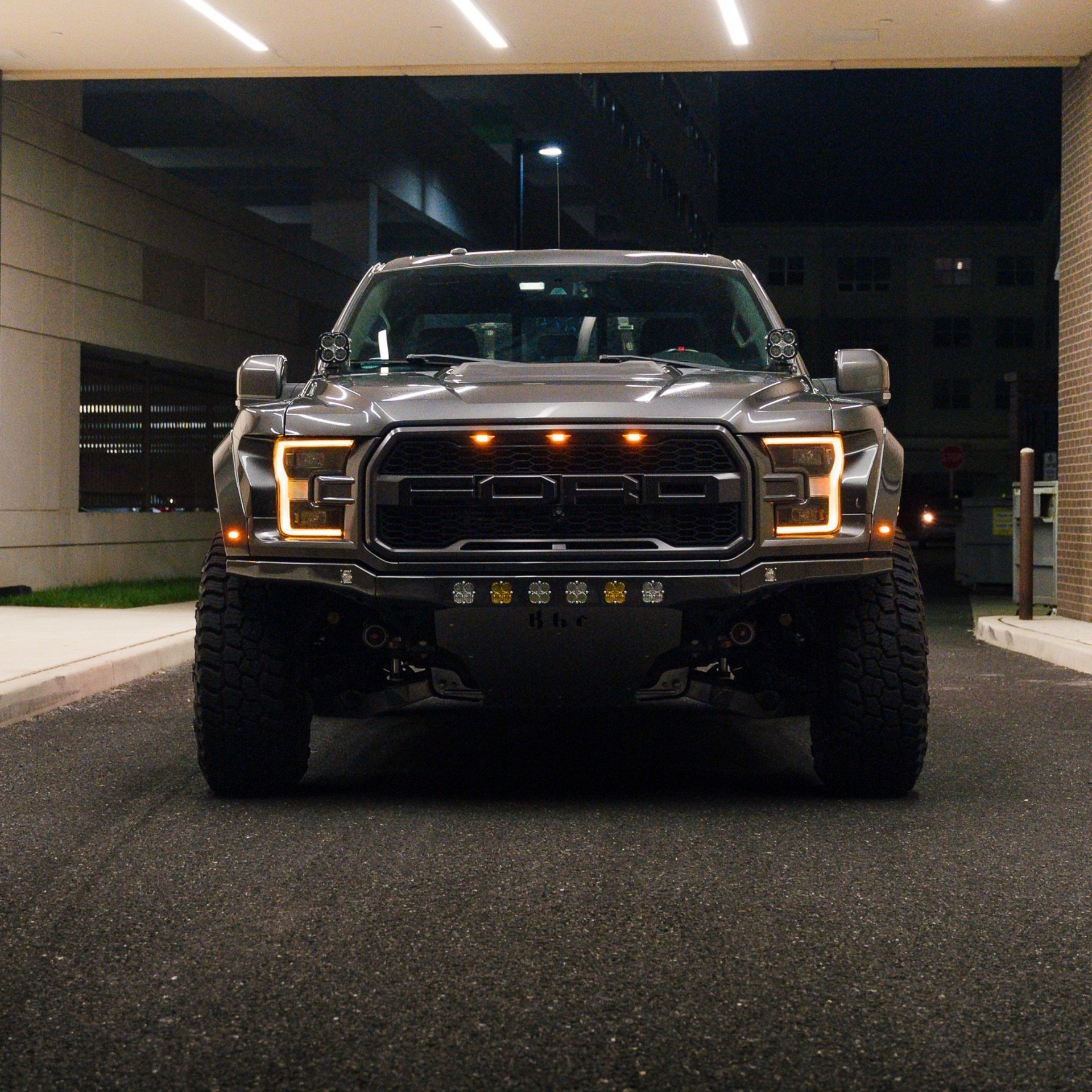 Gray Ford Raptor truck, front view, illuminated headlights and auxiliary lights, parked at night.