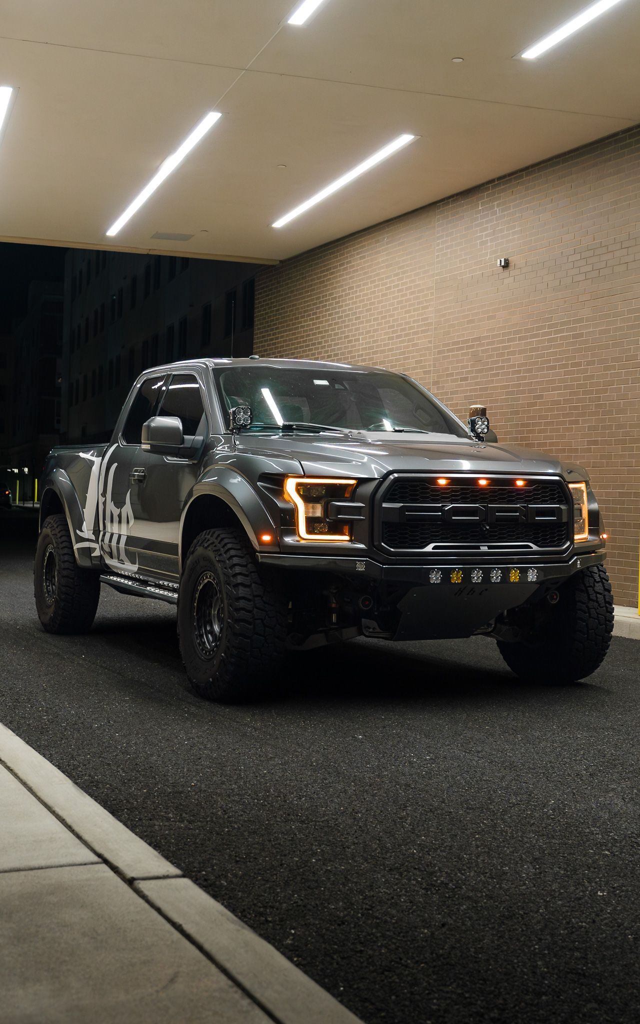 Grey Ford Raptor truck parked under a well-lit awning, large tires and off-road modifications.