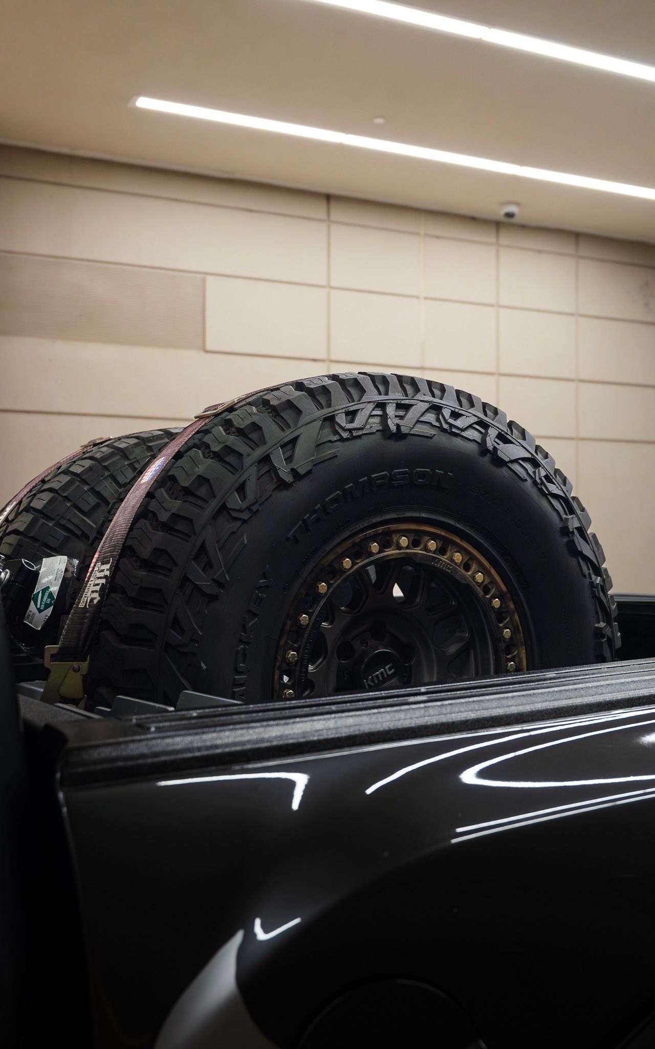 Two large off-road tires secured in a black truck bed. Gold rim detail, tan wall in the background.