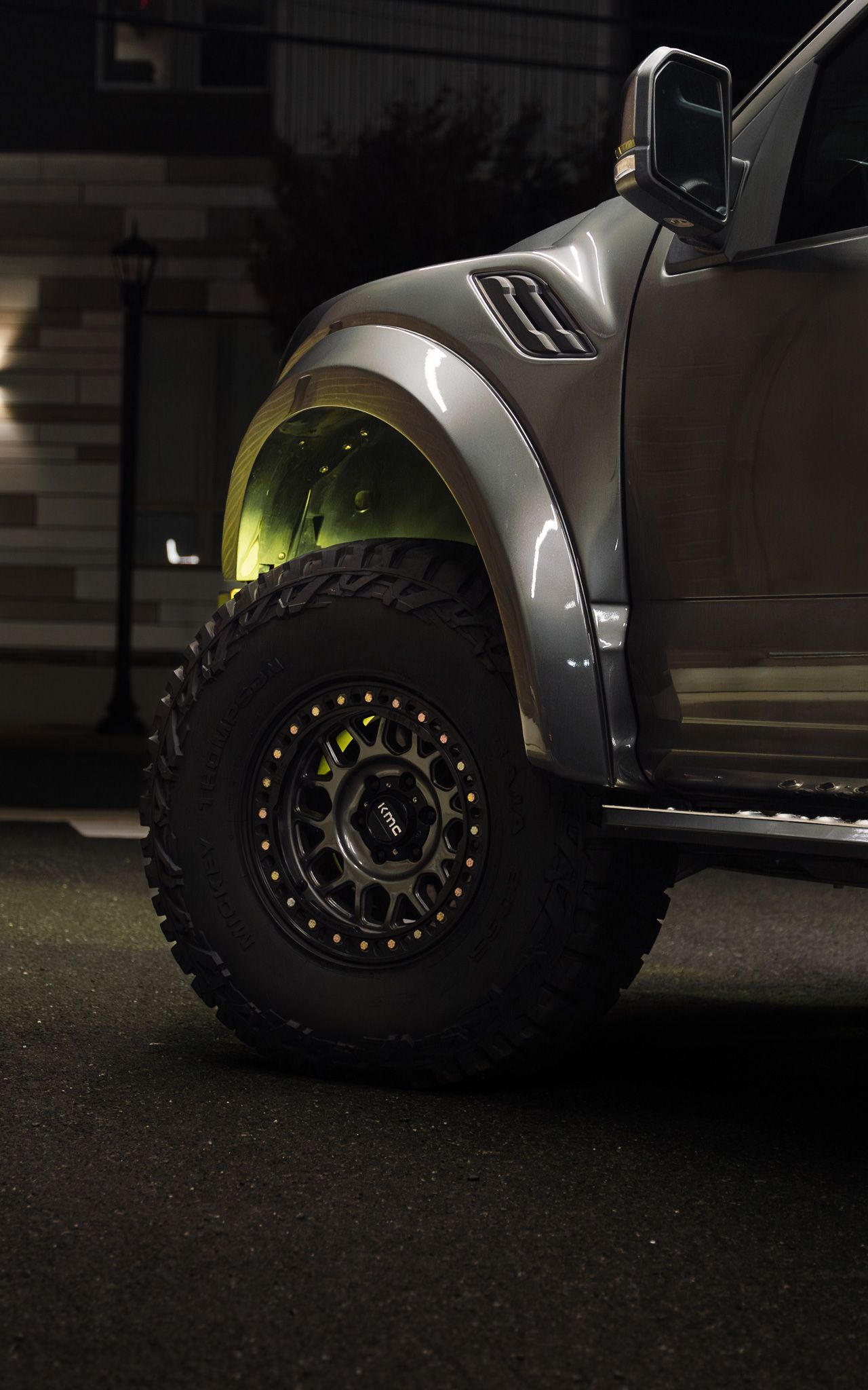 Gray Ford Raptor truck tire and fender with black rim, lit with yellow glow, in a night setting.