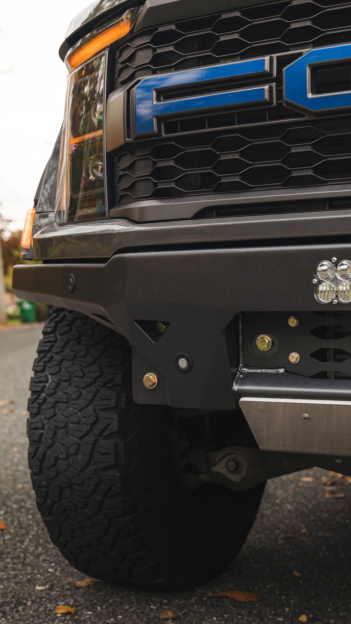 Black off-road truck bumper and tire. The grill has blue lettering, a black bumper and a light bar.