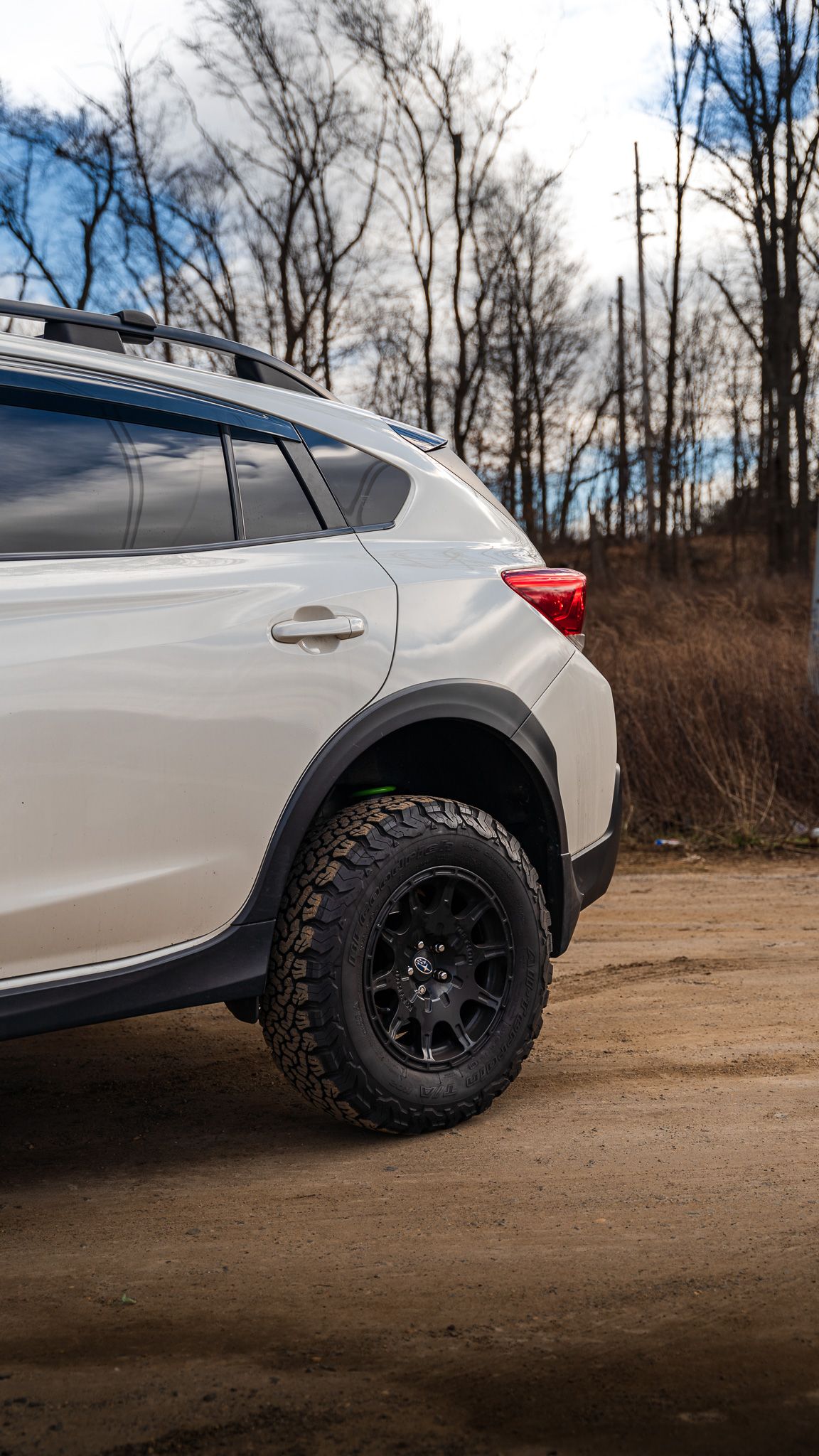 White Subaru Crosstrek with black off-road tires, parked on a dirt road, trees in the background.