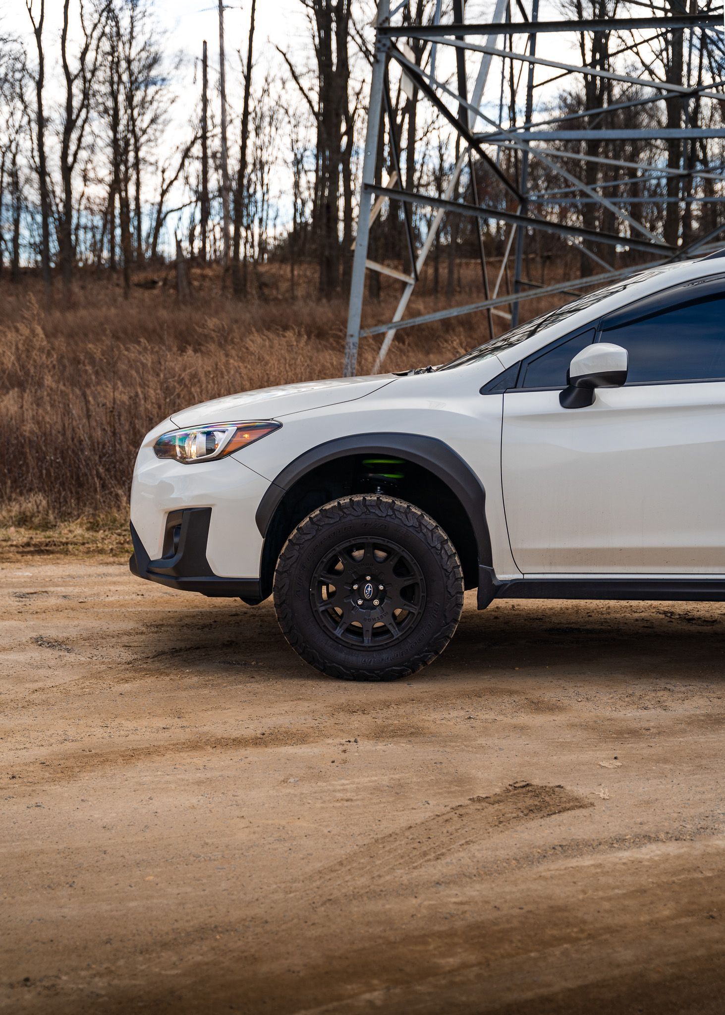 White Subaru Crosstrek with black wheels on a dirt road, trees in the background.