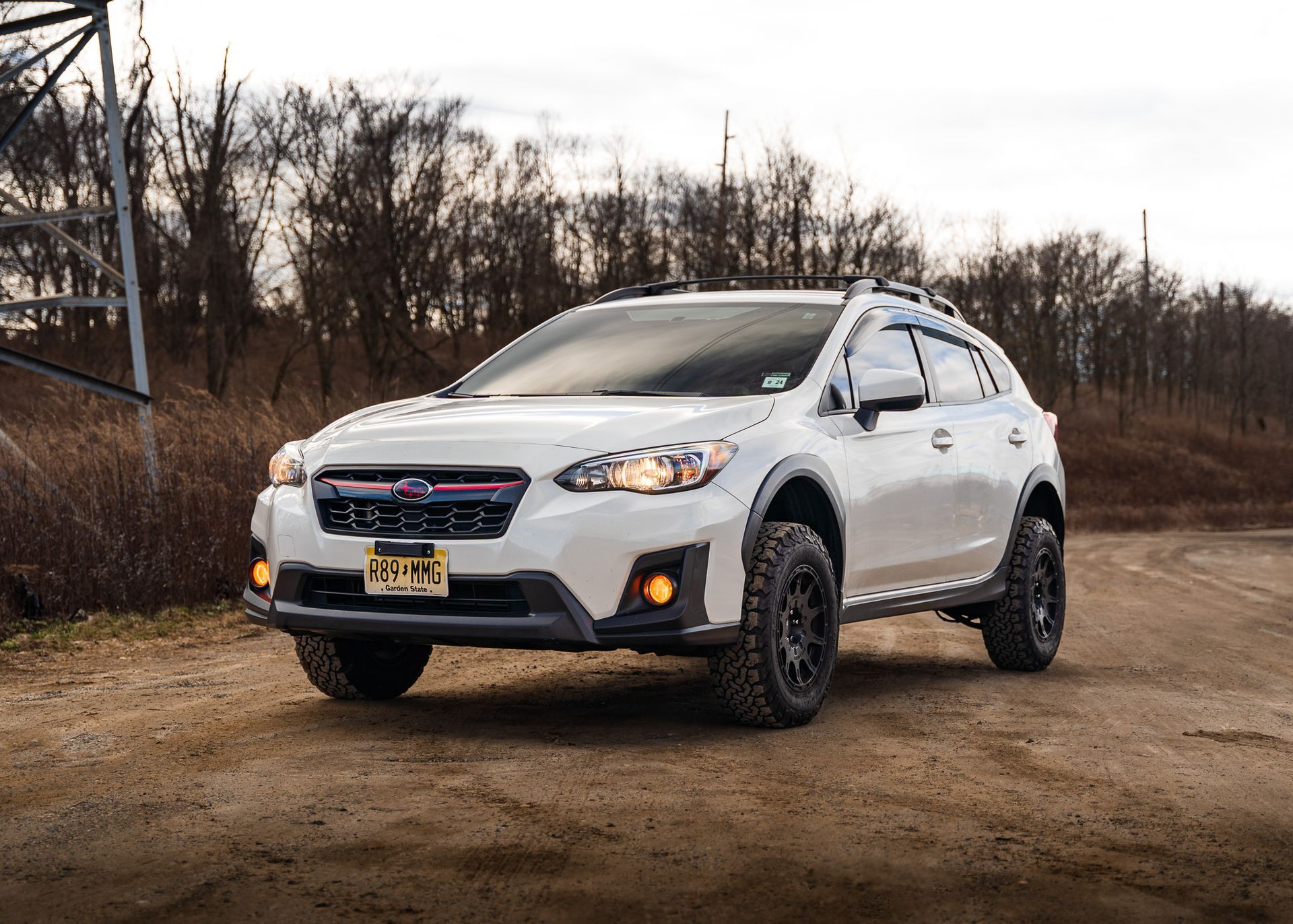 White Subaru Crosstrek SUV parked on dirt road, in front of a treeline, with a utility pole to the left.