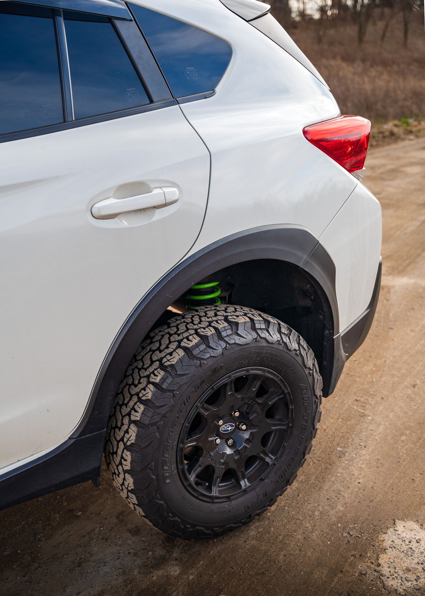 White SUV with black off-road tires, lifted suspension, and green coil spring on a dirt road.