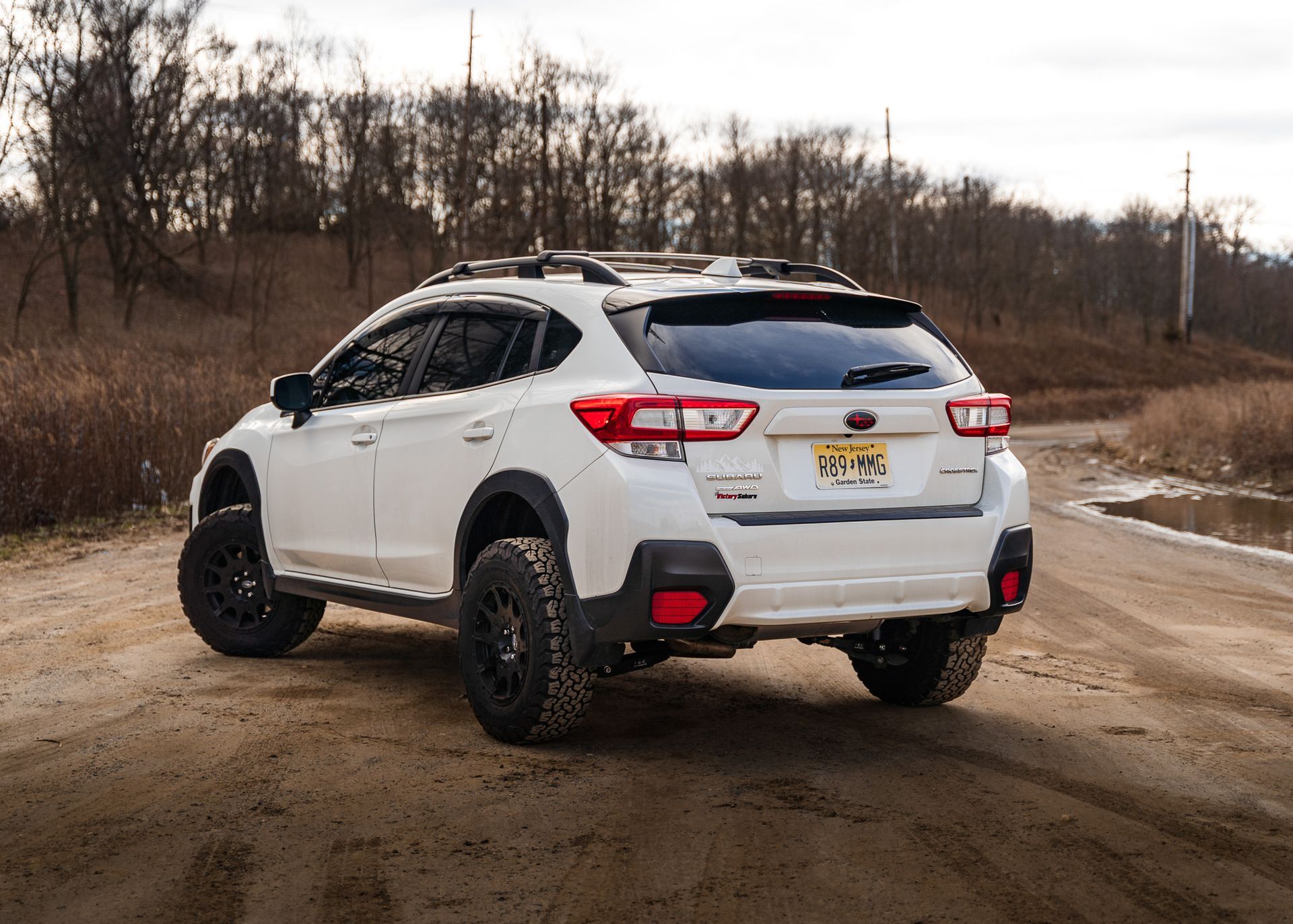 White Subaru Crosstrek on a dirt road, modified with black wheels and a roof rack; wooded background.