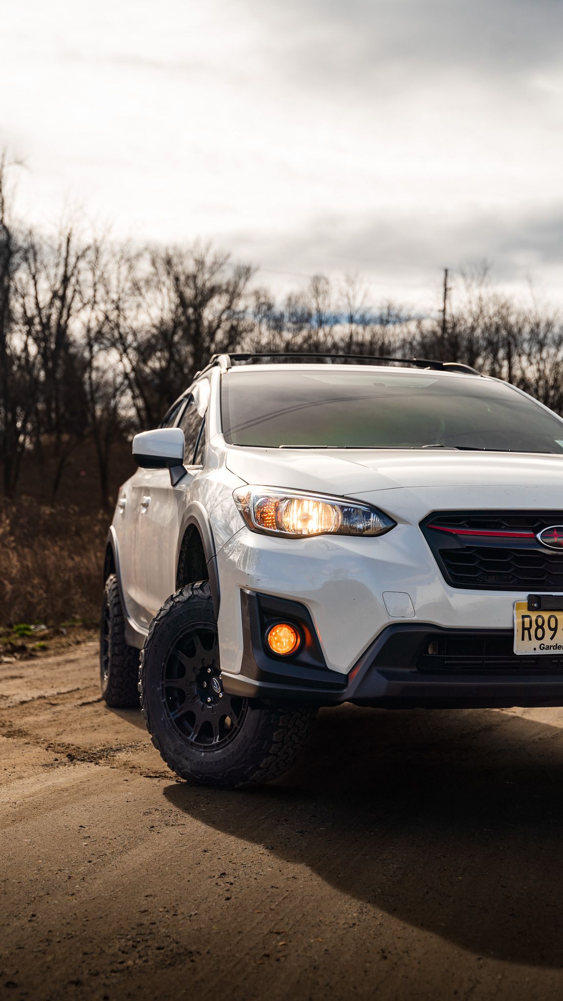 White Subaru Crosstrek on a dirt road. Black wheels, orange fog lights, cloudy sky in background.