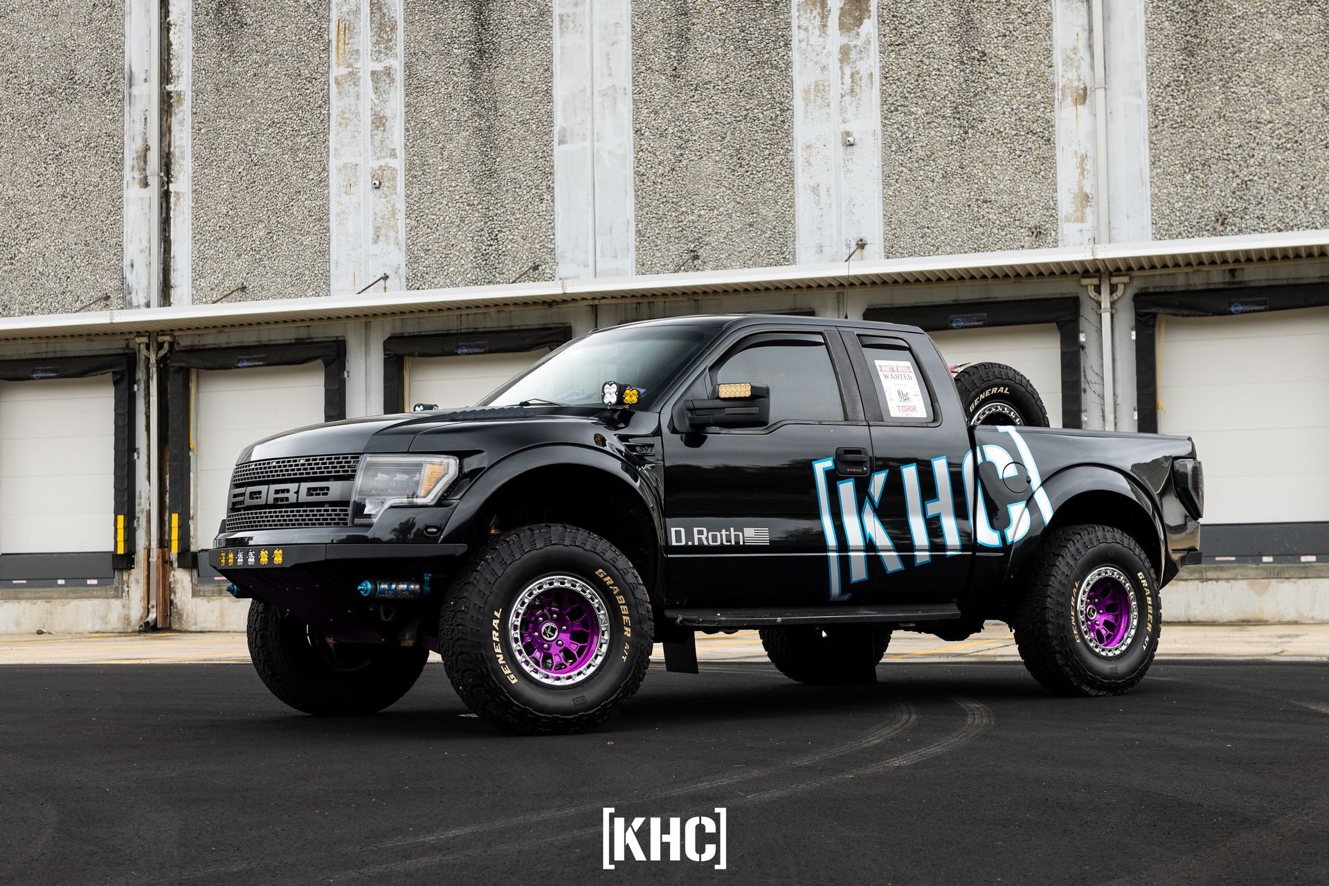 Black Ford Raptor with purple rims and KHC logo, parked in front of a loading dock.