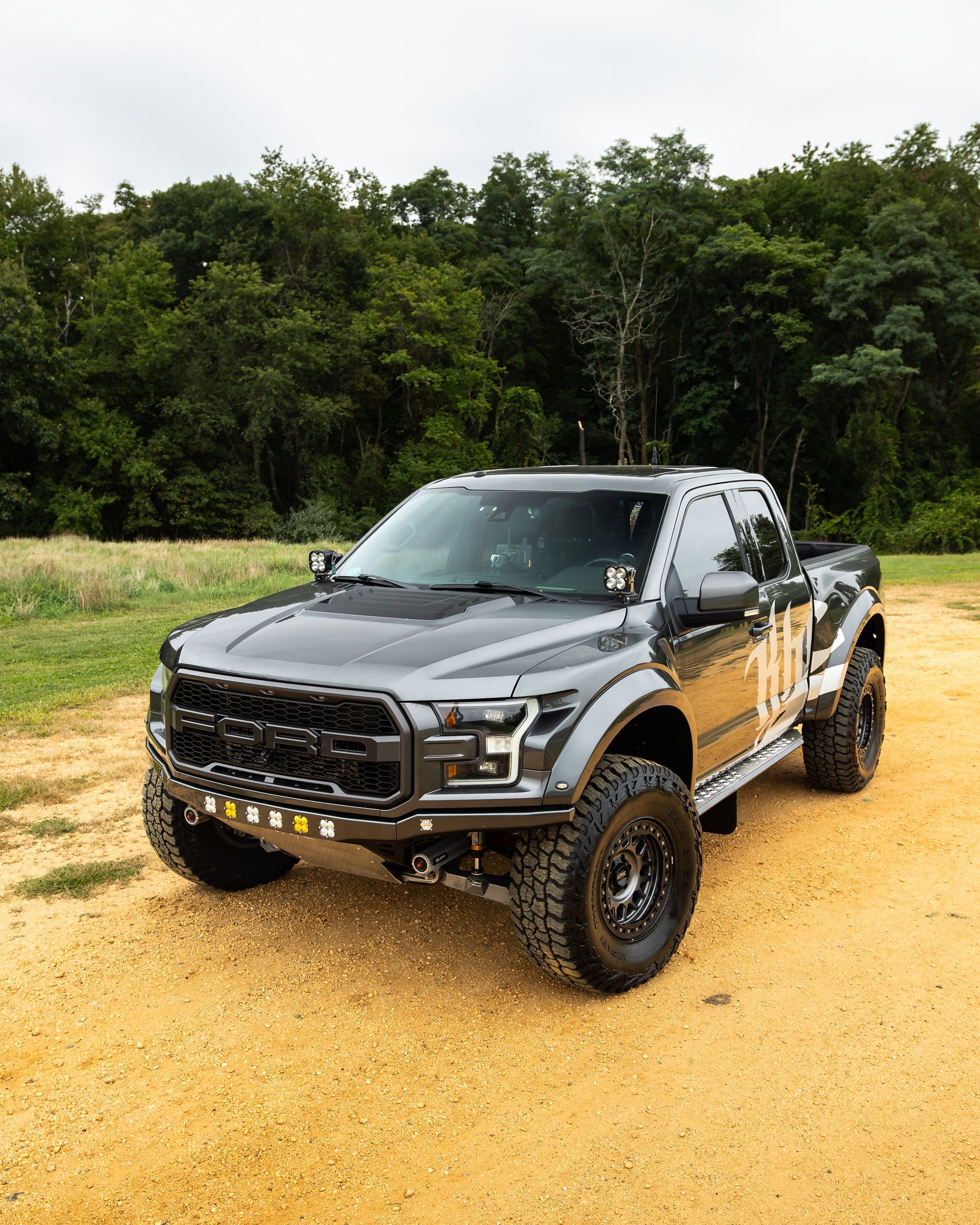 Gray Ford Raptor truck parked on dirt road, surrounded by trees.