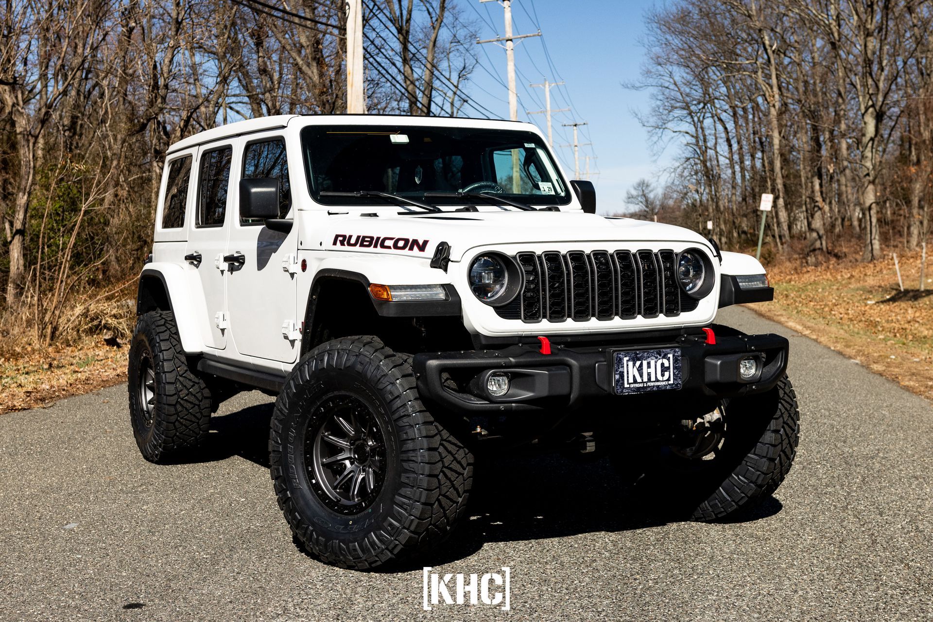 White Jeep Rubicon with large tires parked on a road, trees in the background, sunny day.