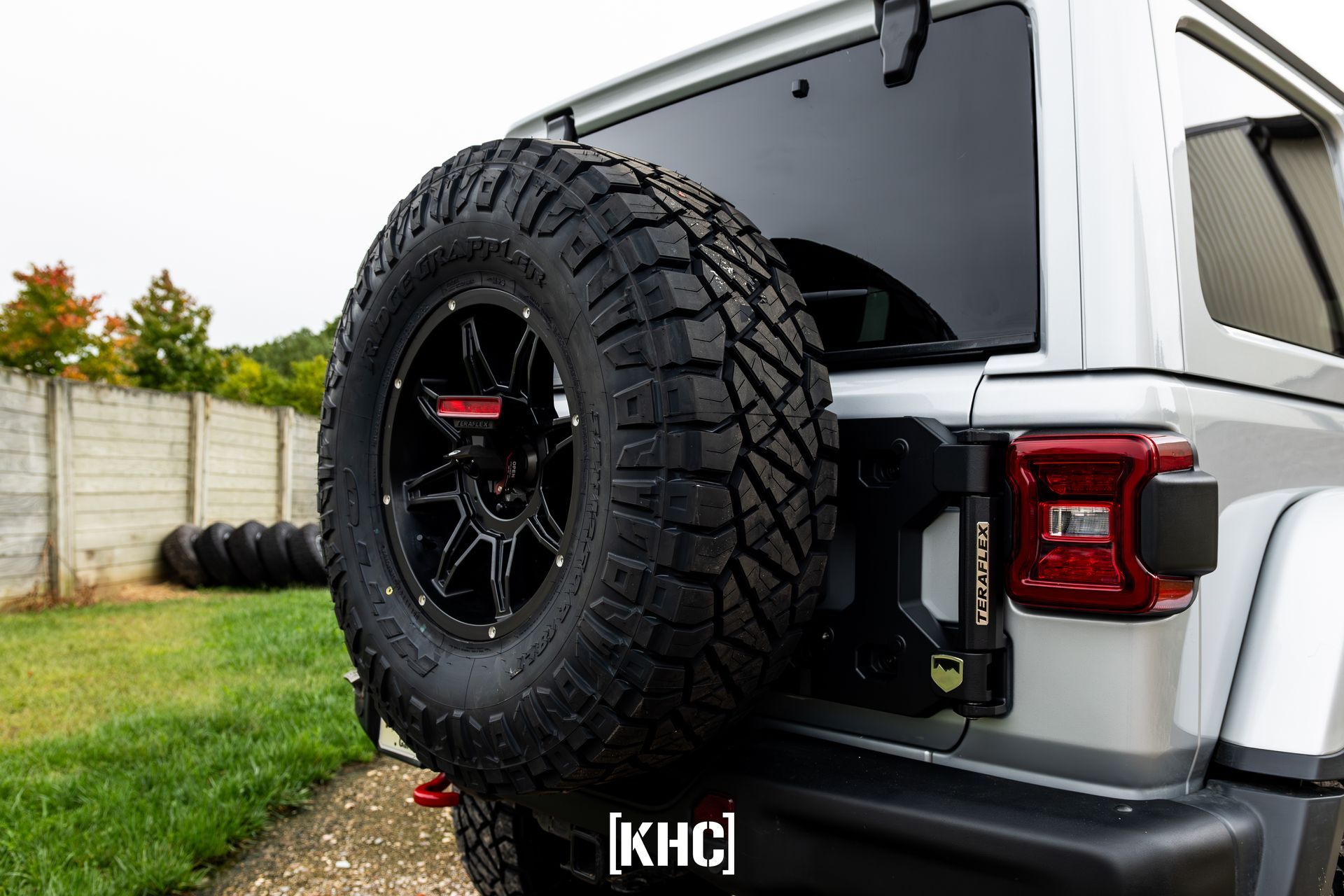 Silver Jeep's rear with a large spare tire mounted on black rim, on a grassy field, next to a concrete wall.