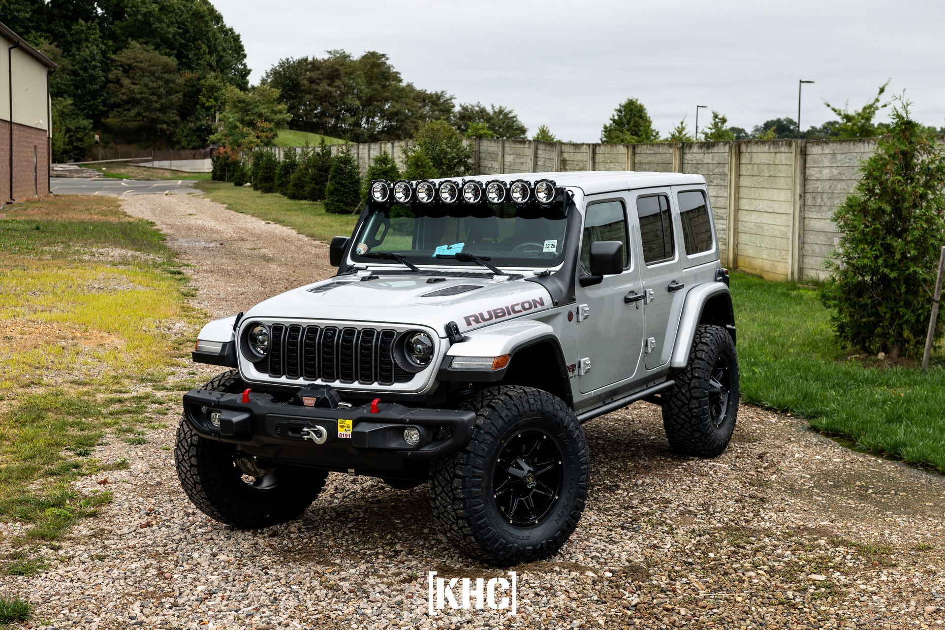 Silver Jeep Wrangler with black accents, light bar, large tires, parked on gravel driveway.