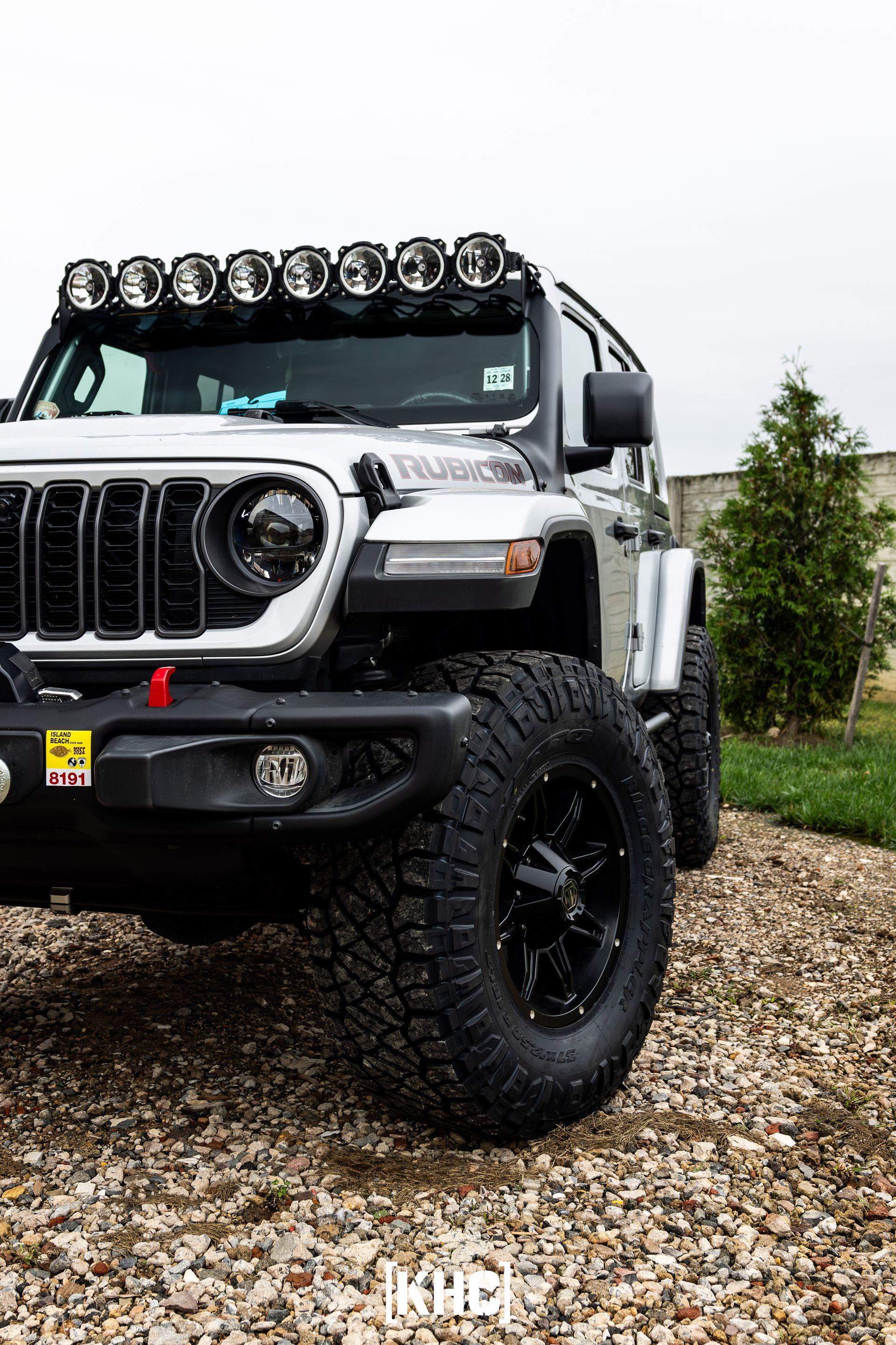 Silver Jeep Wrangler with off-road tires and light bar, parked on gravel.