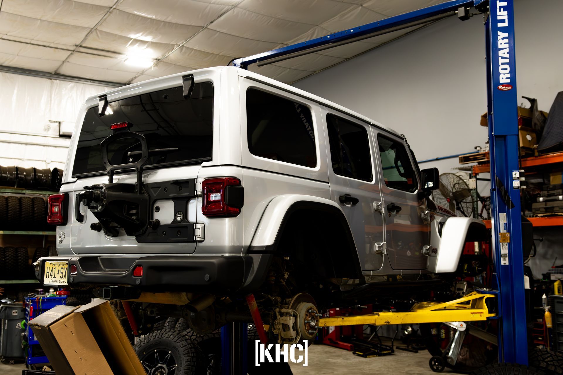 Silver Jeep Wrangler on a hydraulic lift in a repair shop, rear view.