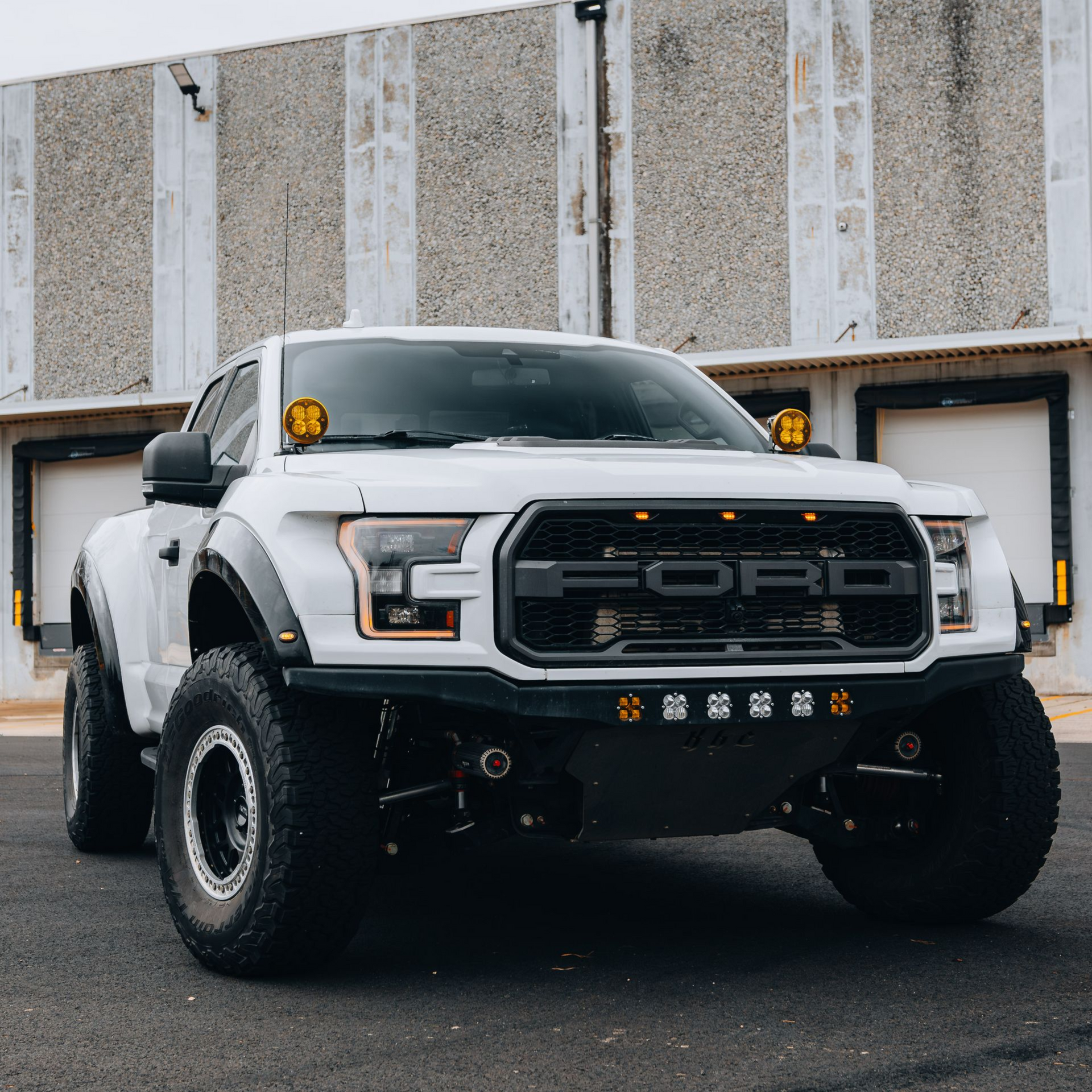 White Ford Raptor off-road truck with black accents and amber lights parked in front of a gray building.
