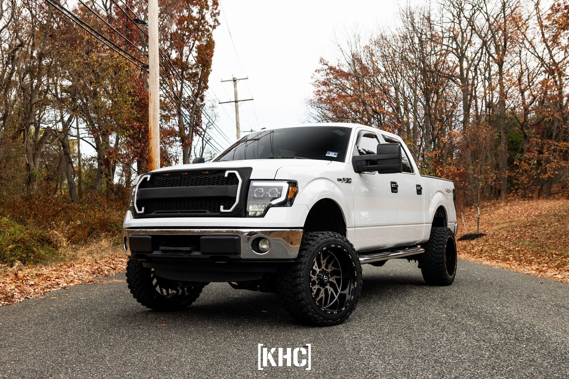 White lifted Ford F-150 truck with black grill and wheels parked on a road, trees in the background.