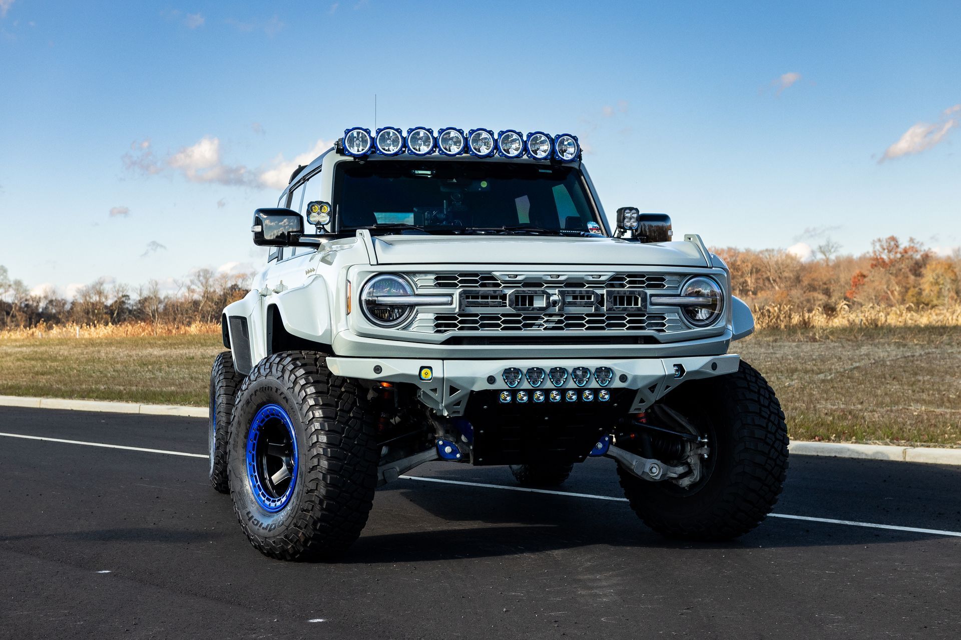 White off-road Ford Bronco with blue accents, large tires, and a light bar, on a paved road.