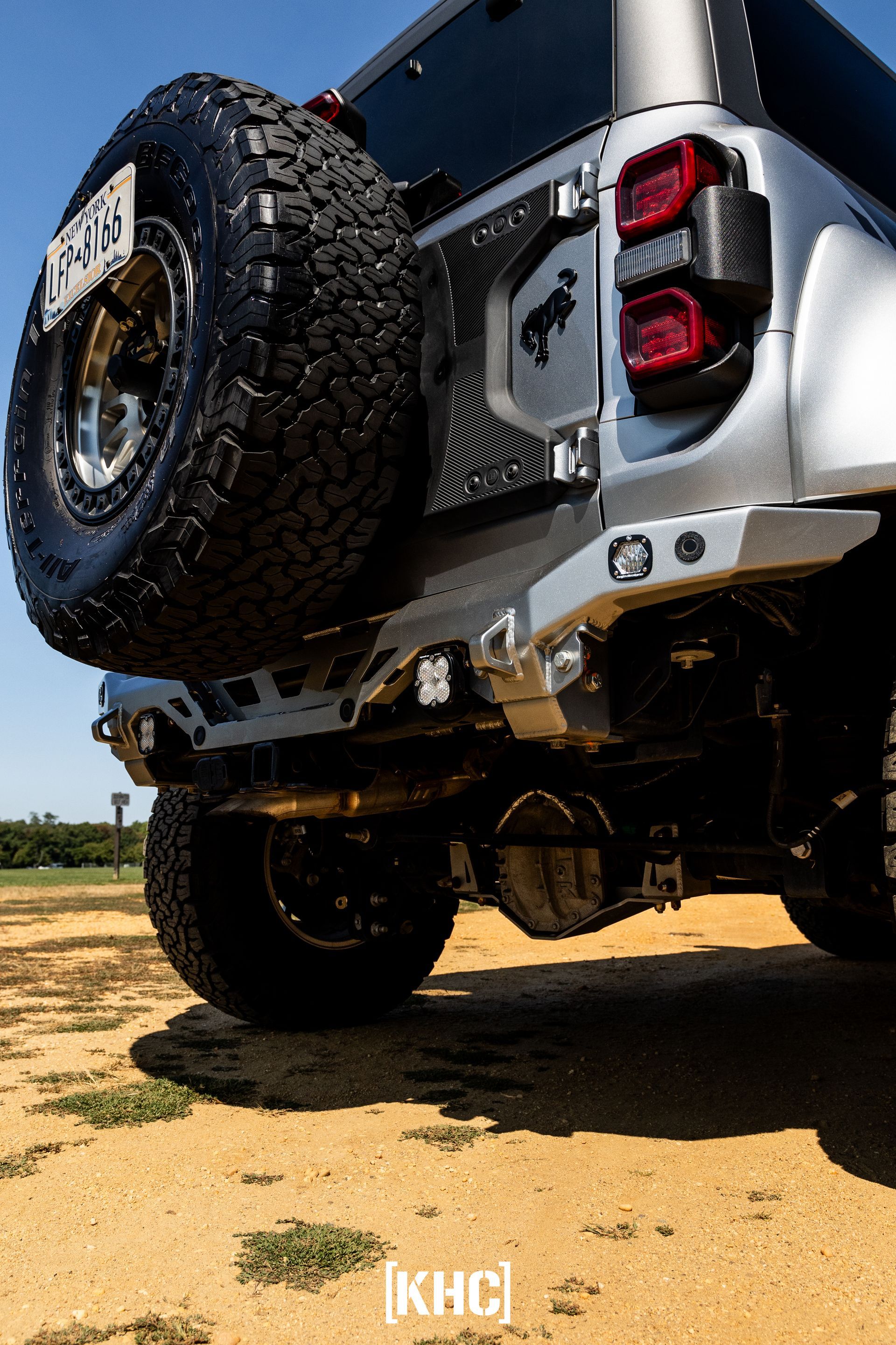 Silver off-road SUV rear with spare tire, modified bumper, black accent, sitting on sand.