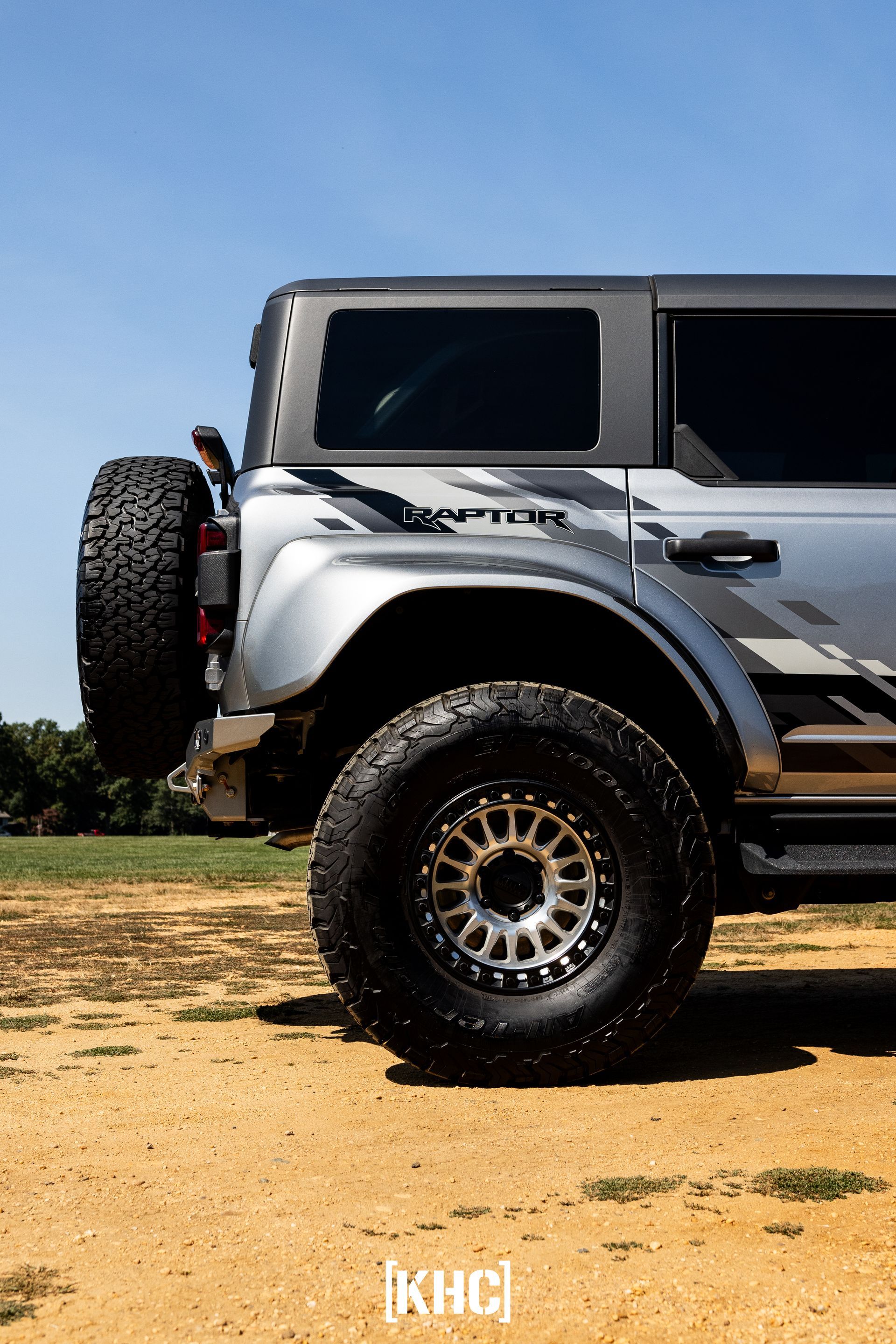 Silver Ford Bronco Raptor, off-road tires, parked on dirt with a blue sky background.