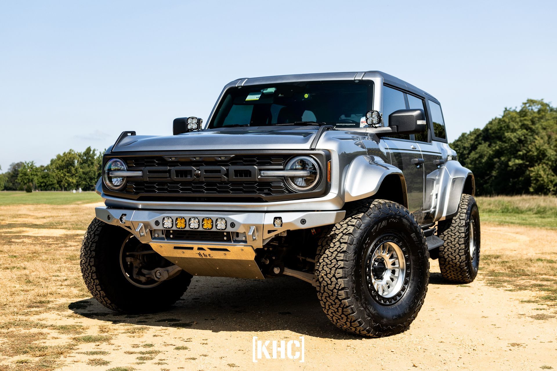 Silver Ford Bronco with large tires and off-road modifications on dirt in a sunny setting.