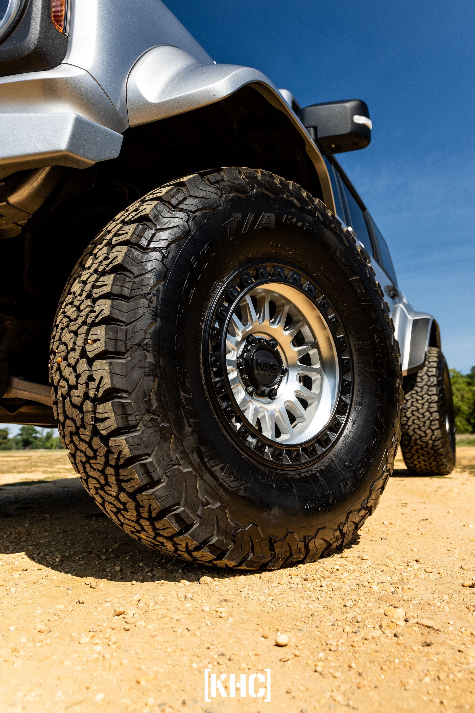Close-up of a silver SUV's off-road tire on a sandy surface, with the blue sky in the background.