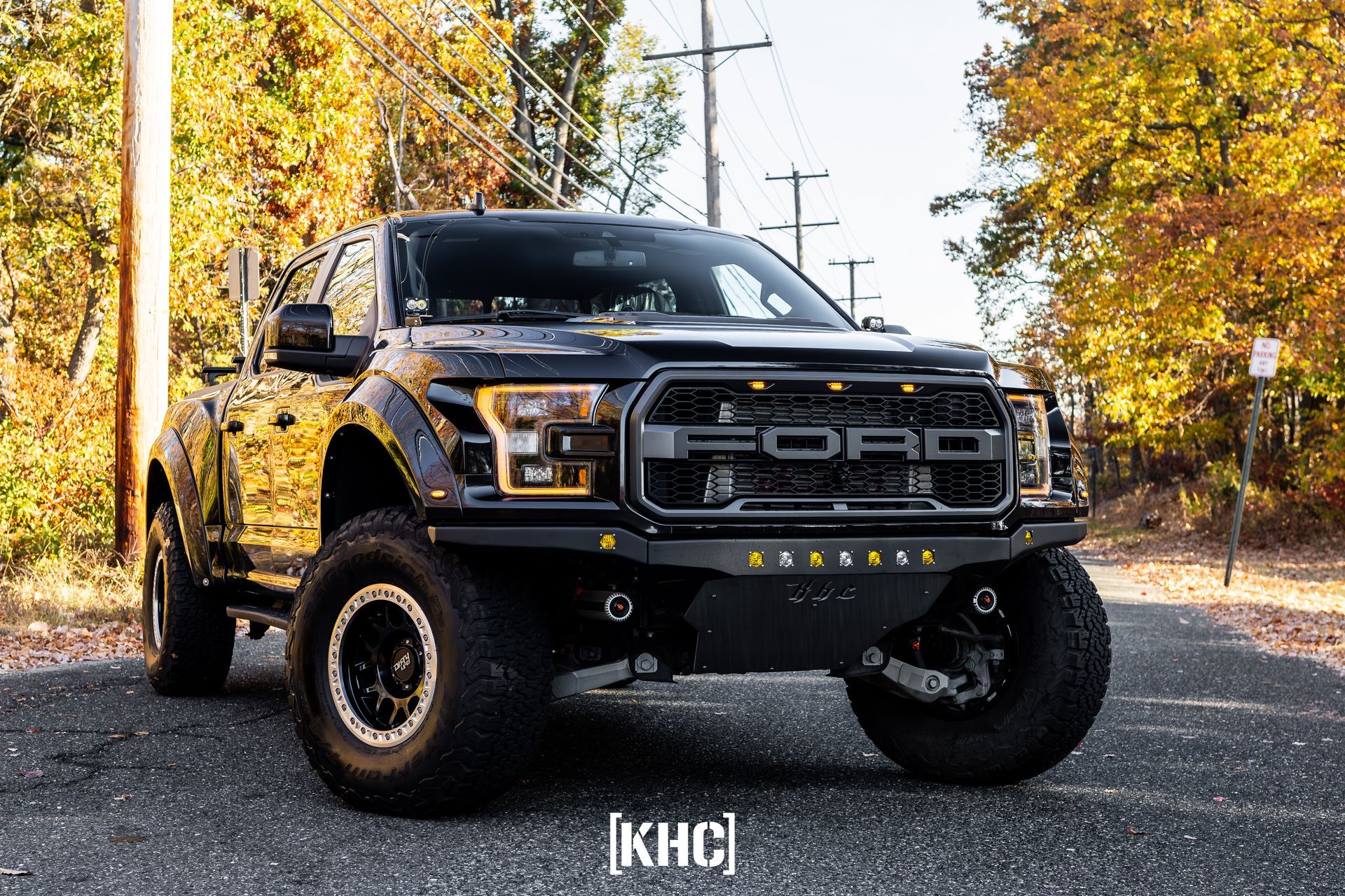 Black Ford Raptor truck on a road with fall foliage. It has an aftermarket front bumper and large tires.