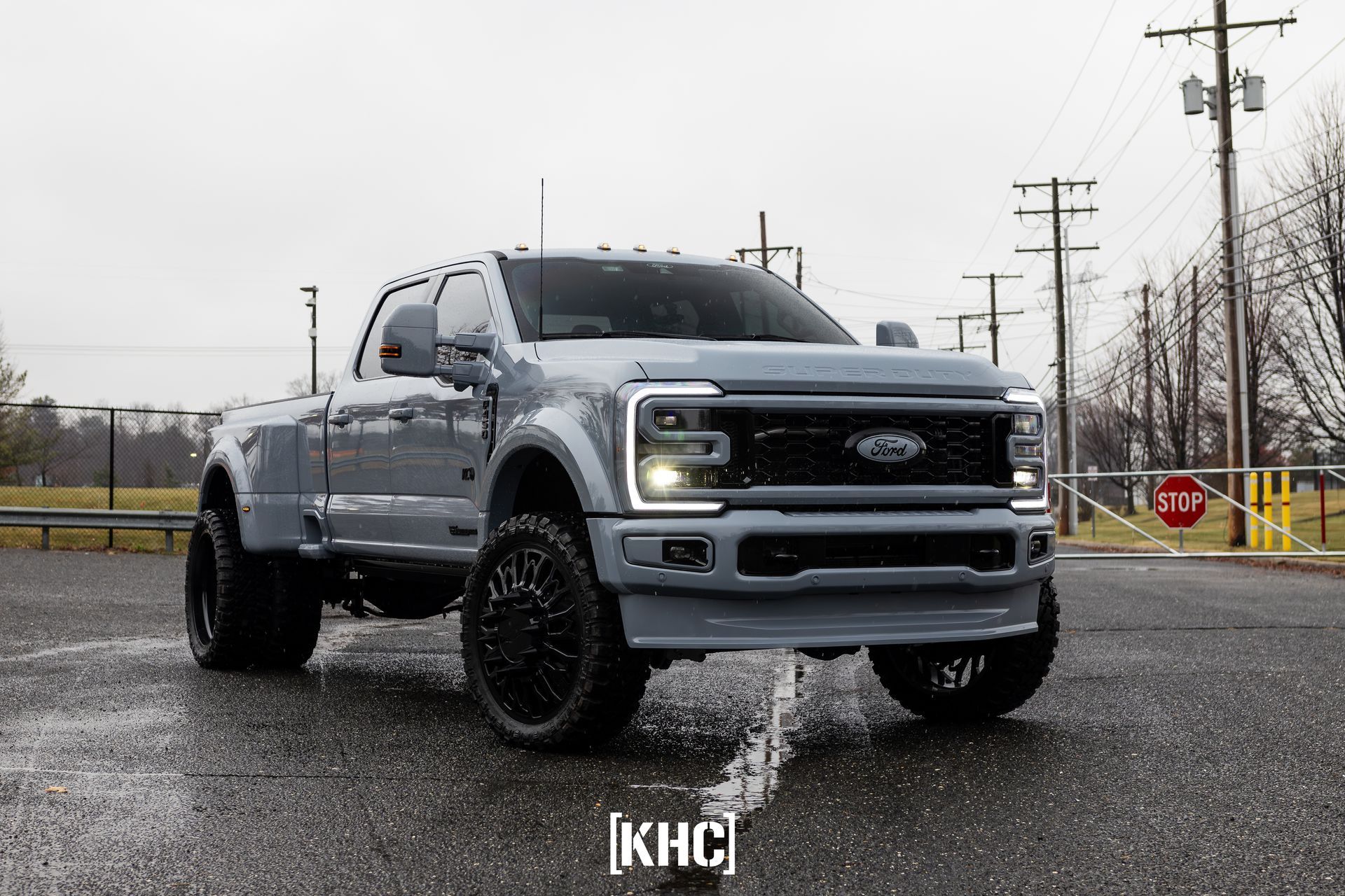 Gray Ford dually pickup truck with black wheels parked on wet pavement in front of a chain-link fence.