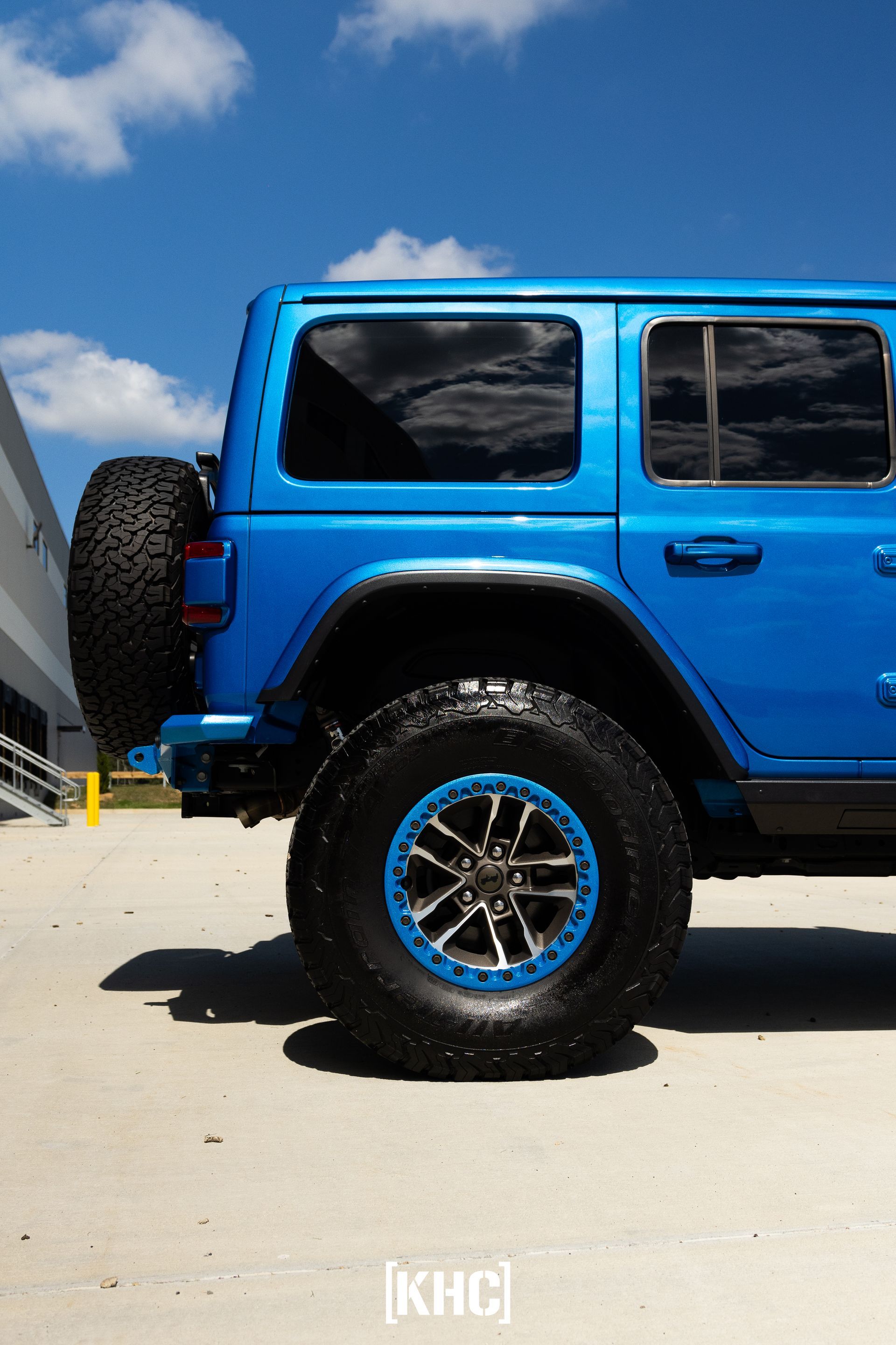 Blue Jeep Wrangler with black tires and blue rims parked outside on a sunny day.