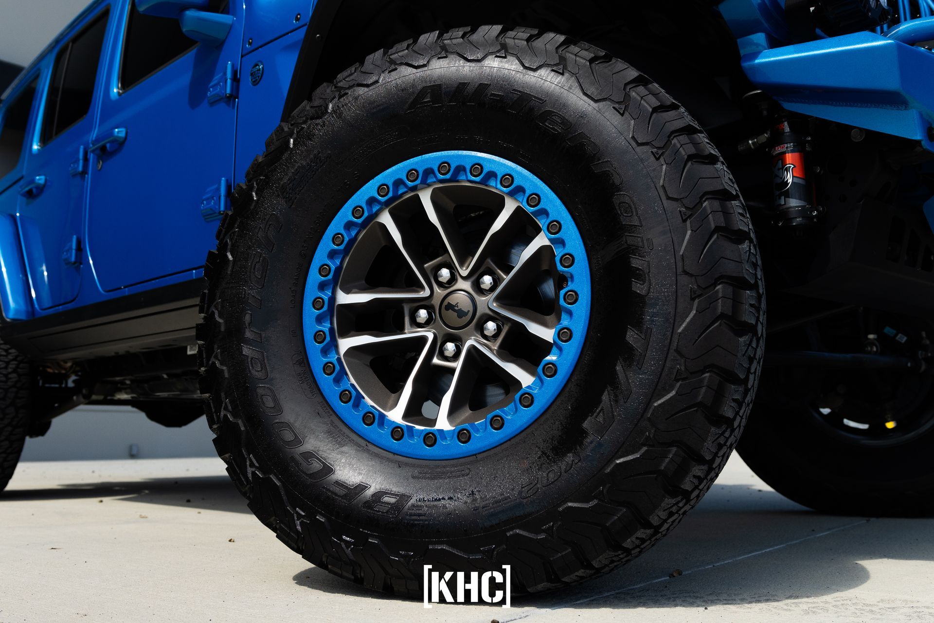Blue Jeep tire with black and blue rim against a light gray surface.