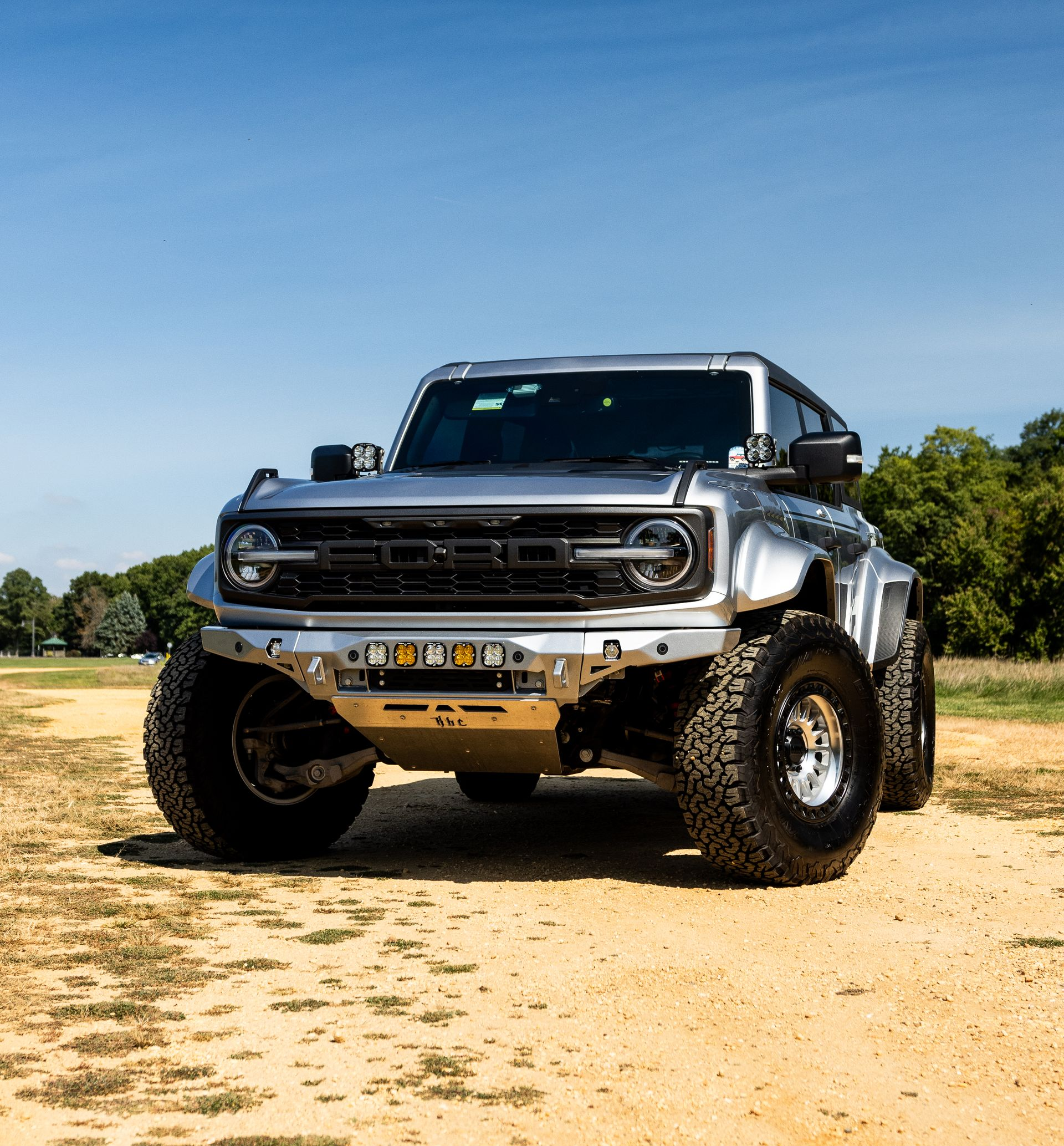 Silver Ford Bronco off-road vehicle with large tires on a dirt road, sunny outdoor setting.