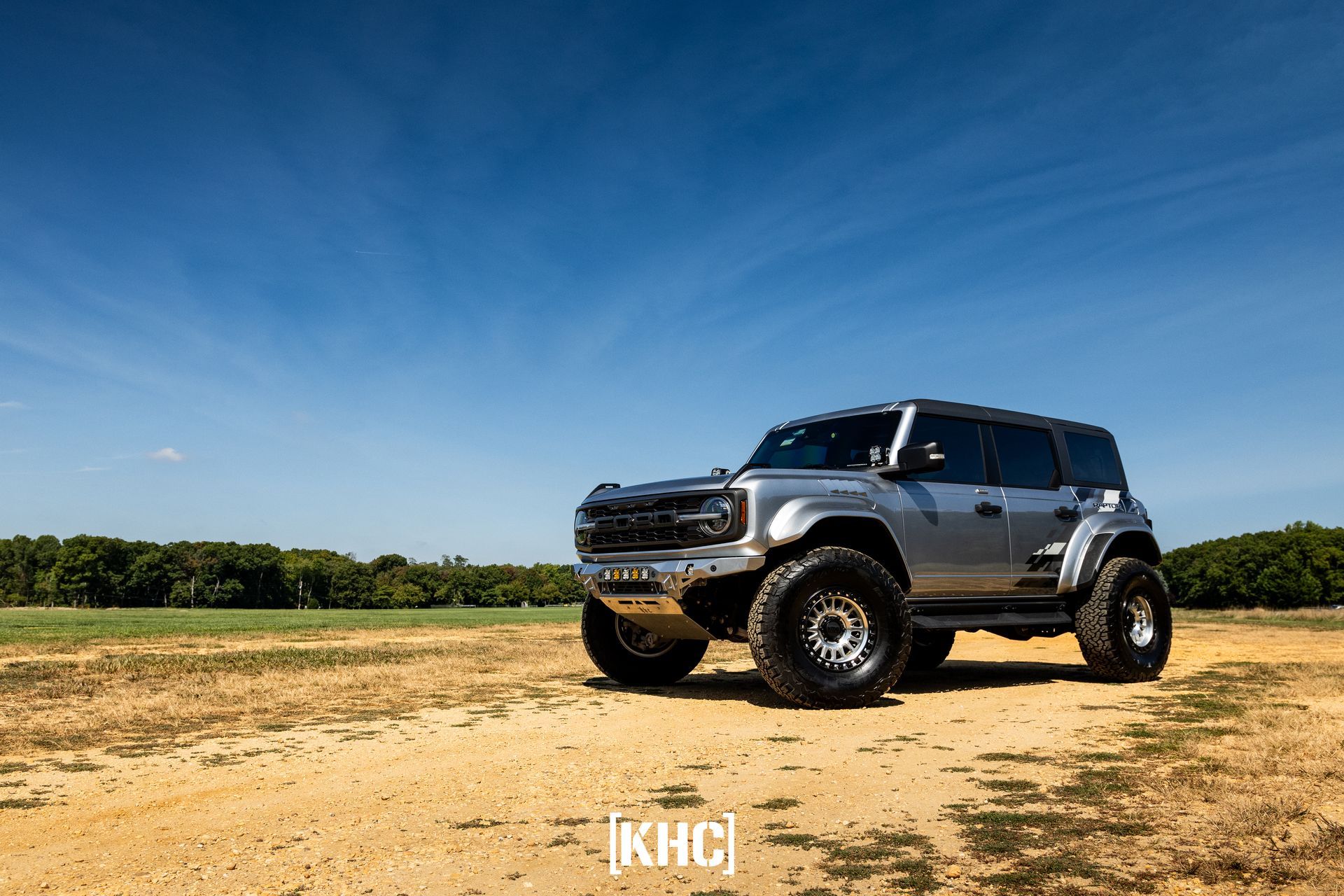 Silver Ford Bronco SUV with large off-road tires, parked on dirt field under blue sky.