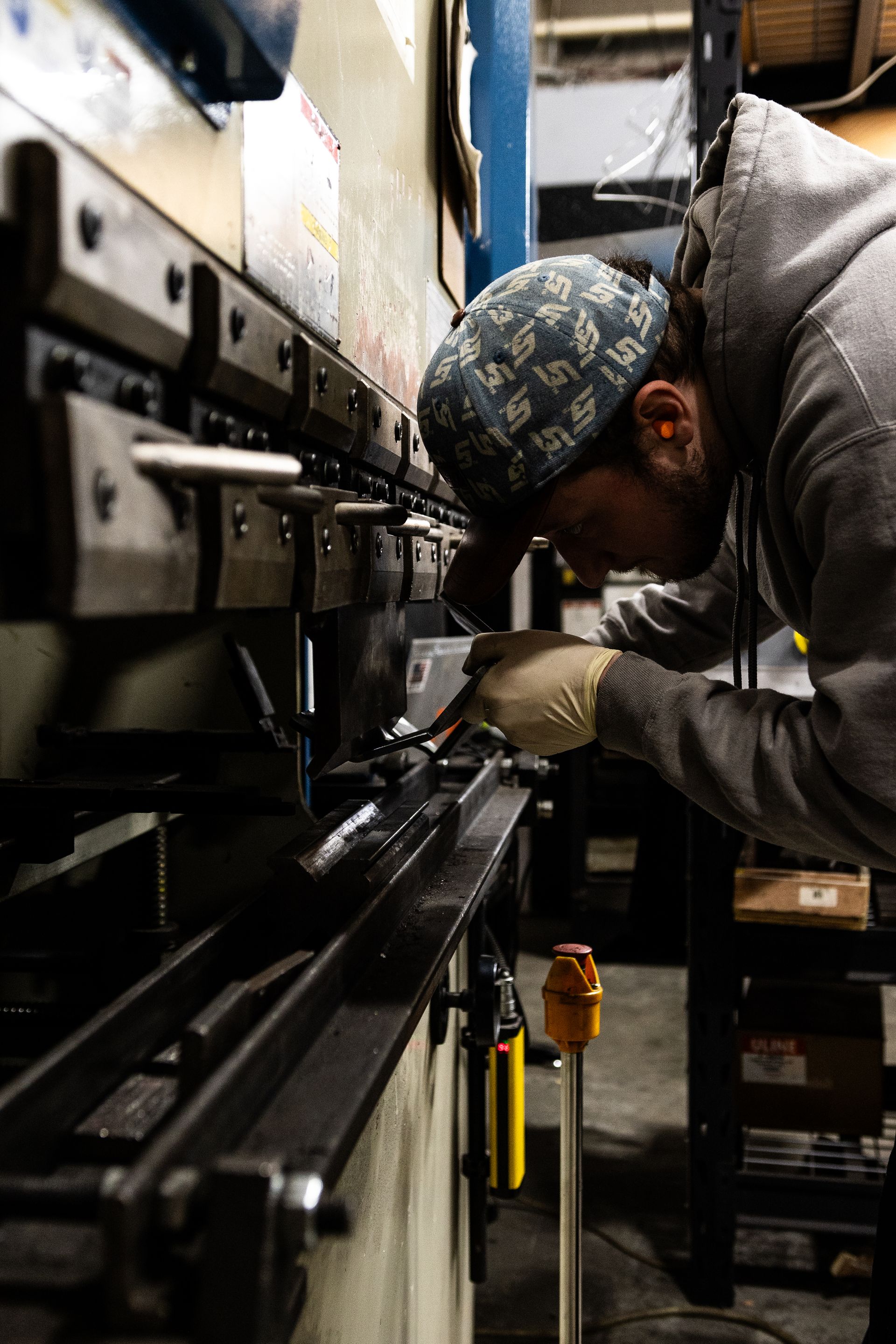Person bends metal sheet on a press brake machine. Factory setting; wearing a hat, gloves, and hoodie.