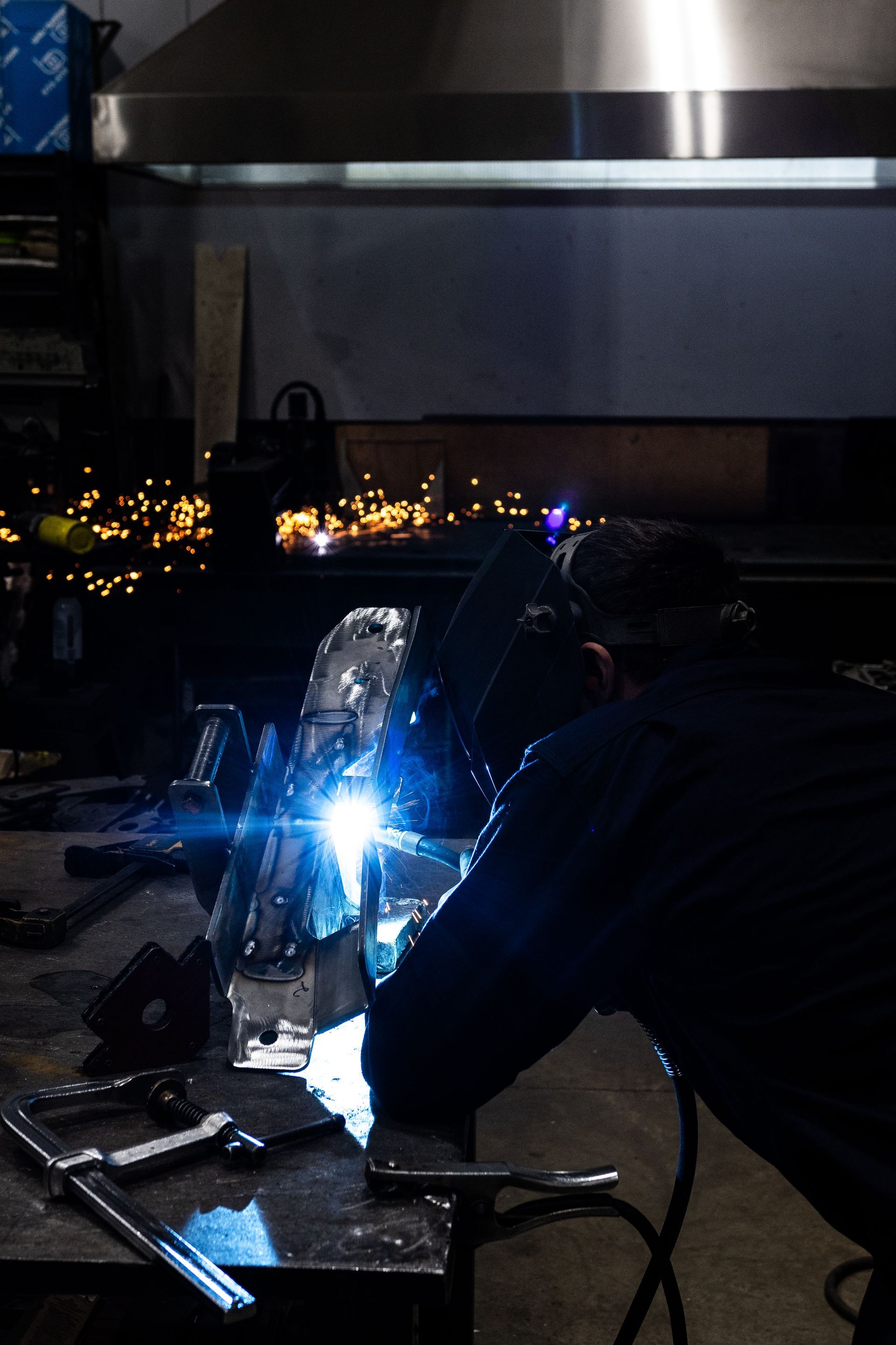 Welder working in a shop, sparks flying from metal, wearing a mask.