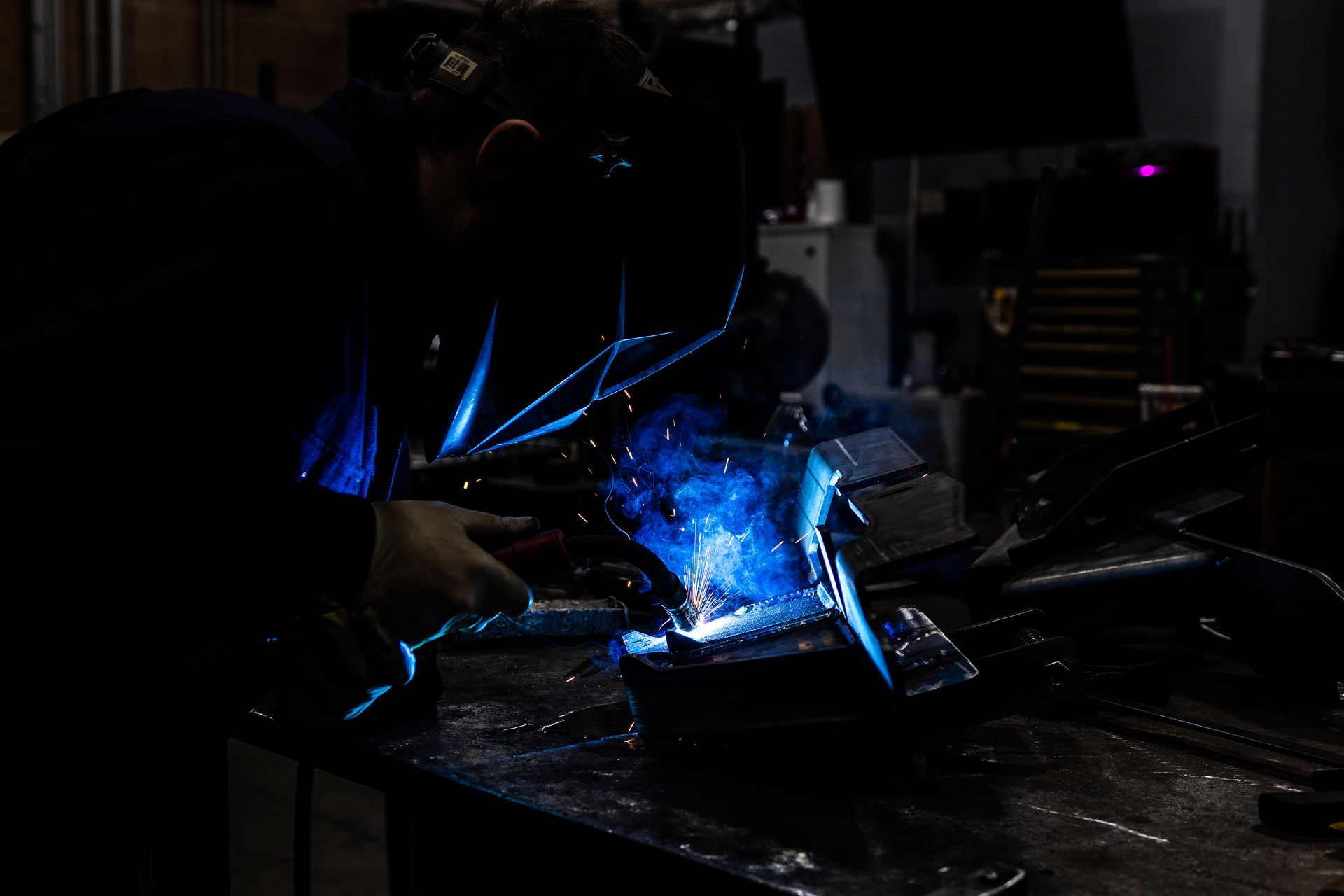 A person welds metal in a dim workshop, with bright blue sparks and light.