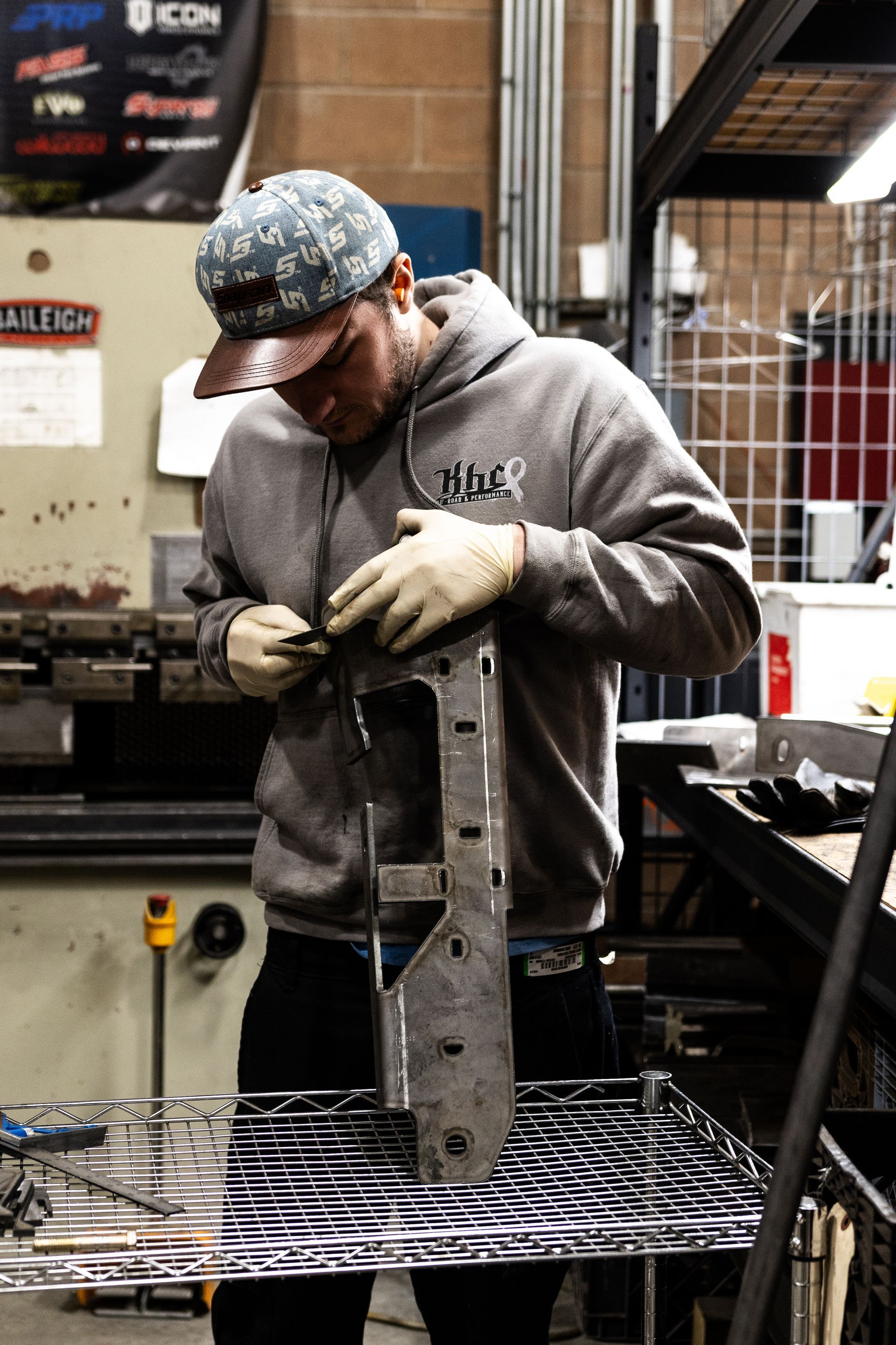 Man in a workshop inspecting metal piece, wearing a gray hoodie and cap.